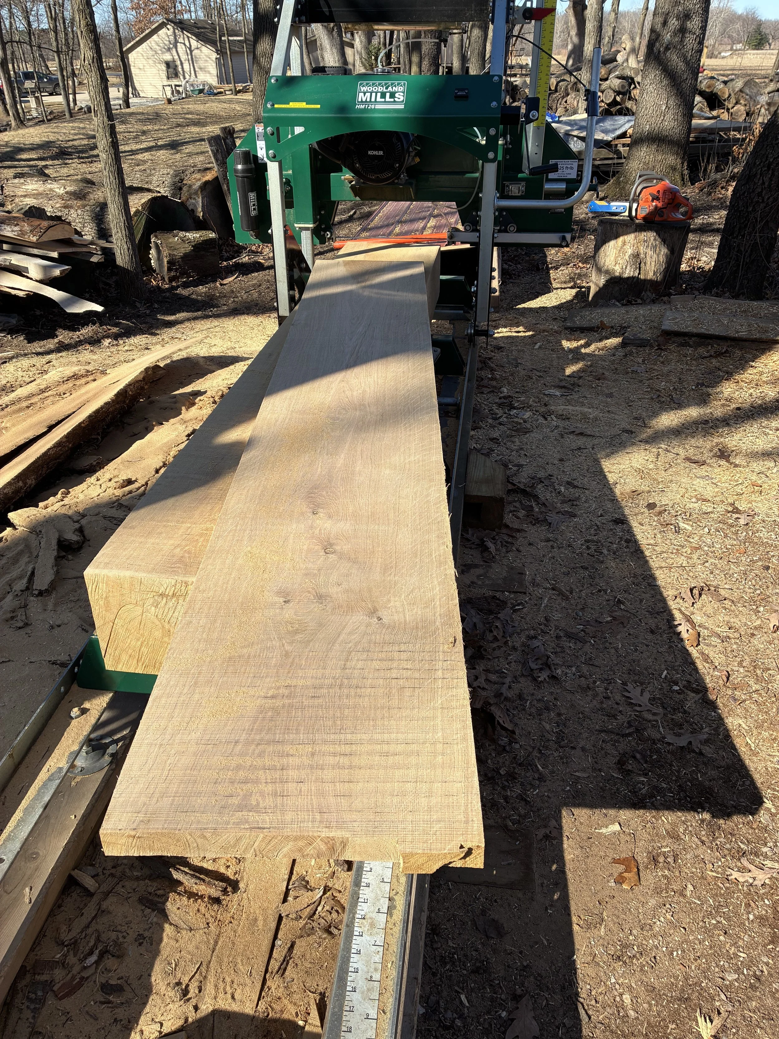A wood planer machine in use outdoors, planing a large piece of wood, with a yard and trees in the background.