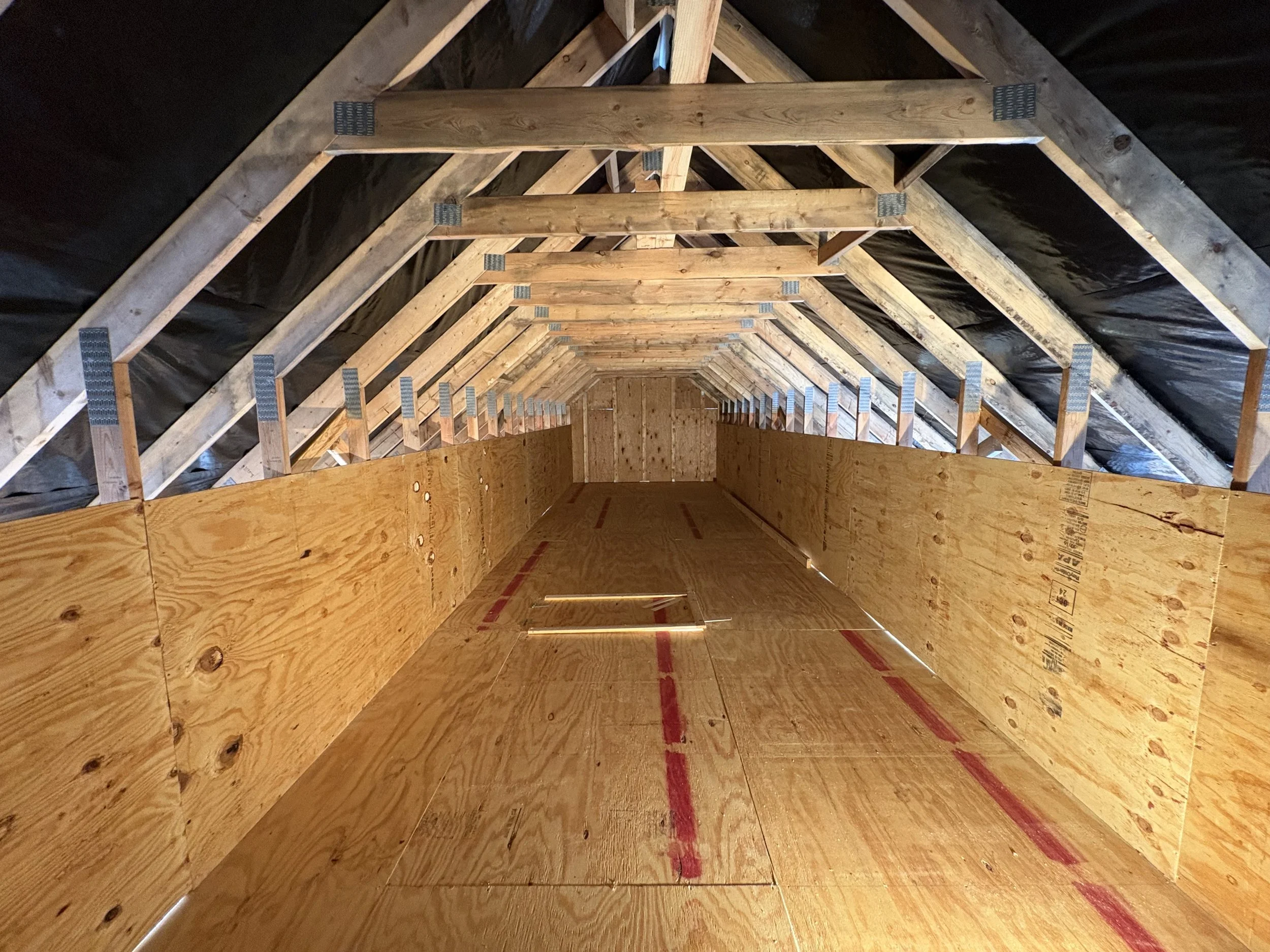 Interior view of an attic under construction, showing wooden framing, plywood walls and floors, and black insulation on the roof.