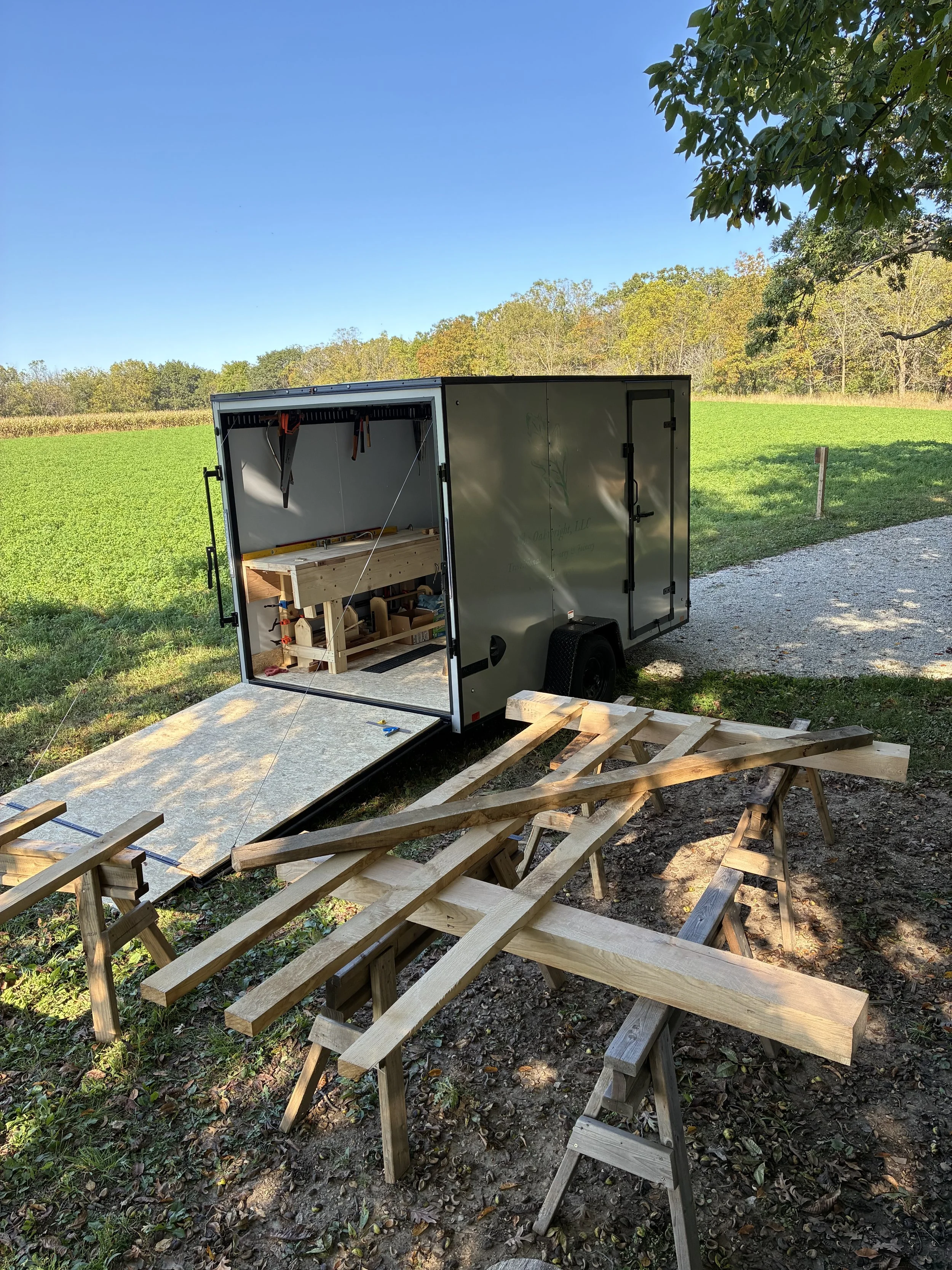 A portable workshop trailer with a ramp open, set up in a grassy area with various wooden planks and workbenches for woodworking projects.