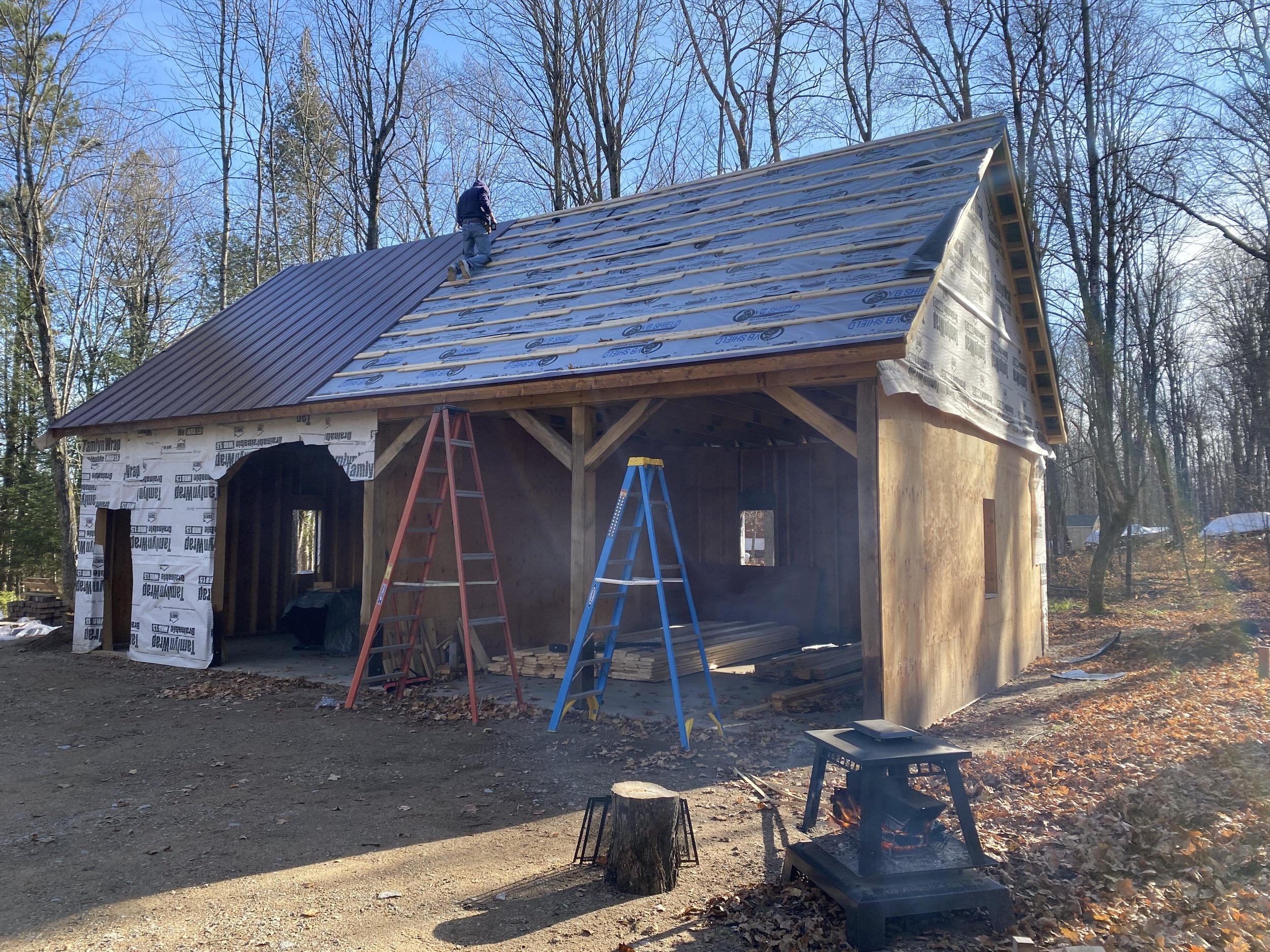 A house under construction with partially installed roofing materials, scaffolding, ladders, and tools visible, surrounded by trees on a clear day.