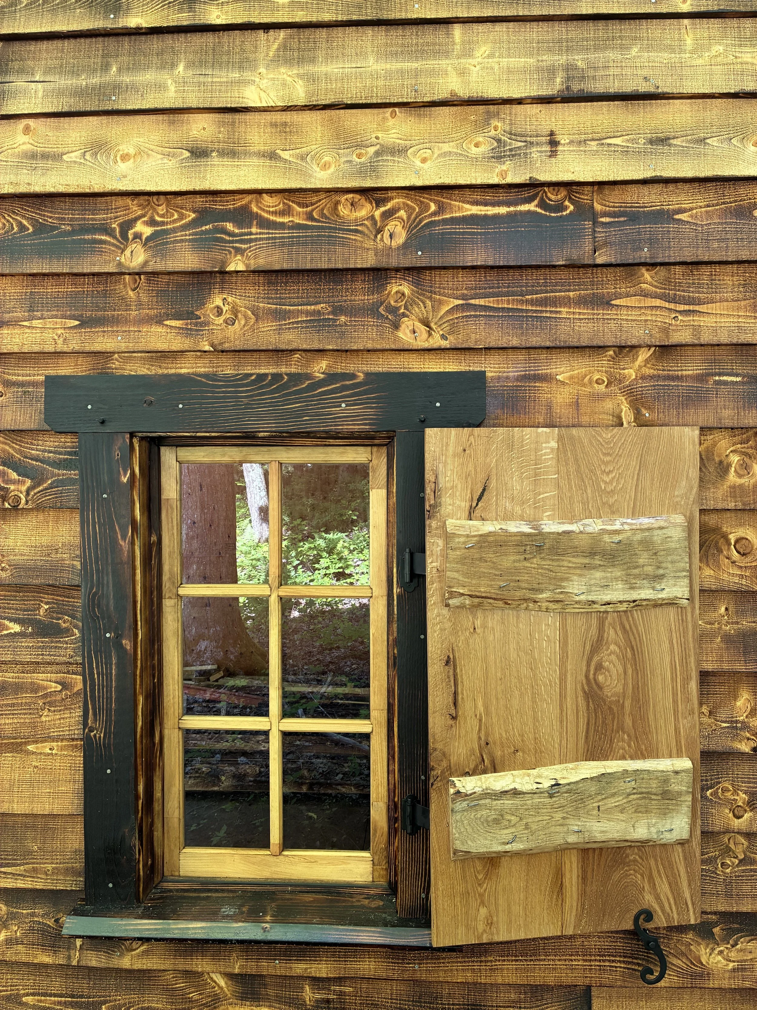 Close-up of a small wooden window with a window shutter, surrounded by a dark-stained wooden frame, on a rustic wooden wall.