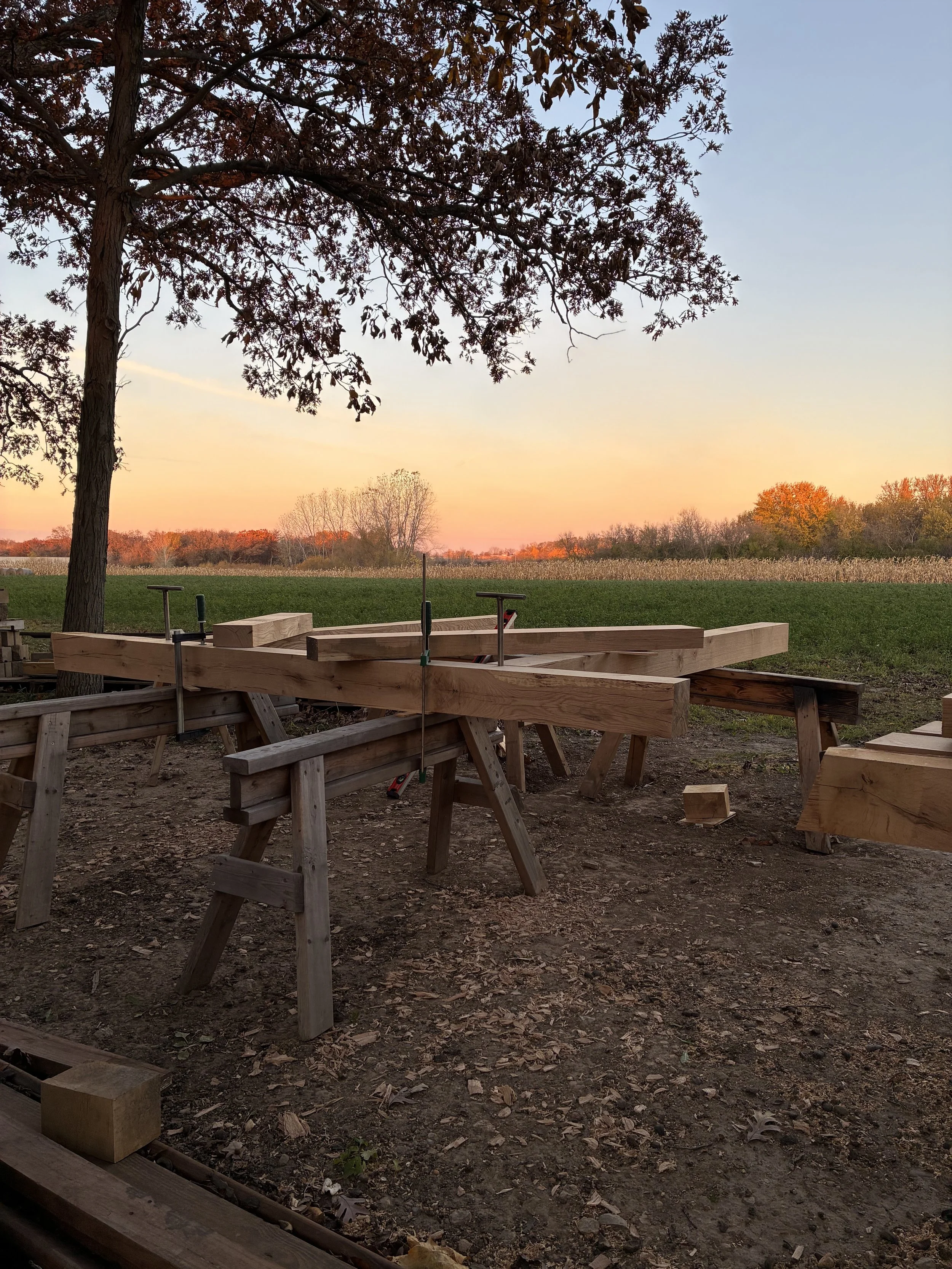 Wooden construction work in progress outdoors during sunset, with trees and a field in the background.