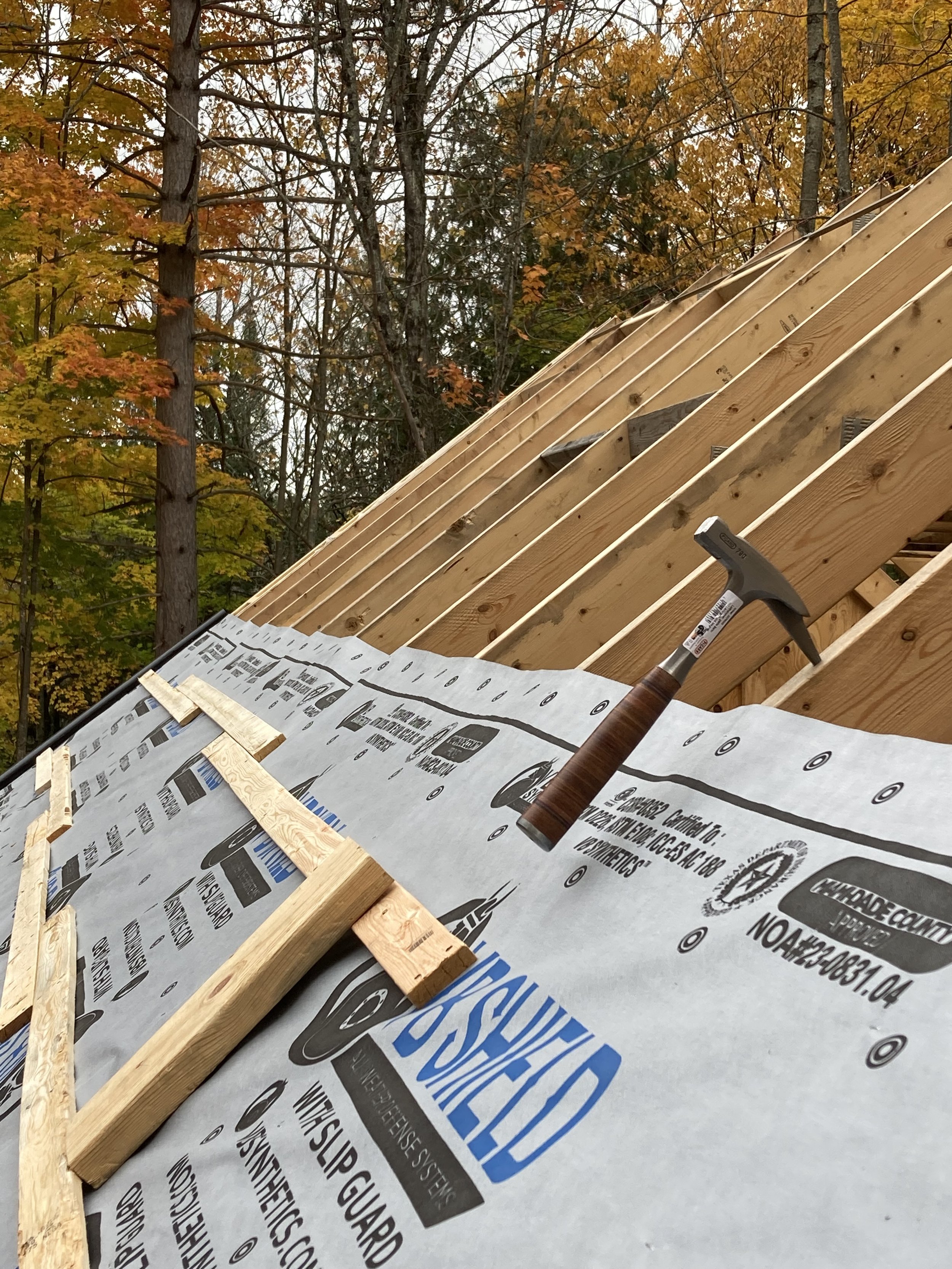A construction site showing the framing of a roof with wooden beams, a hammer resting on the beams, and building wrap partially installed with yellow and orange autumn trees in the background.