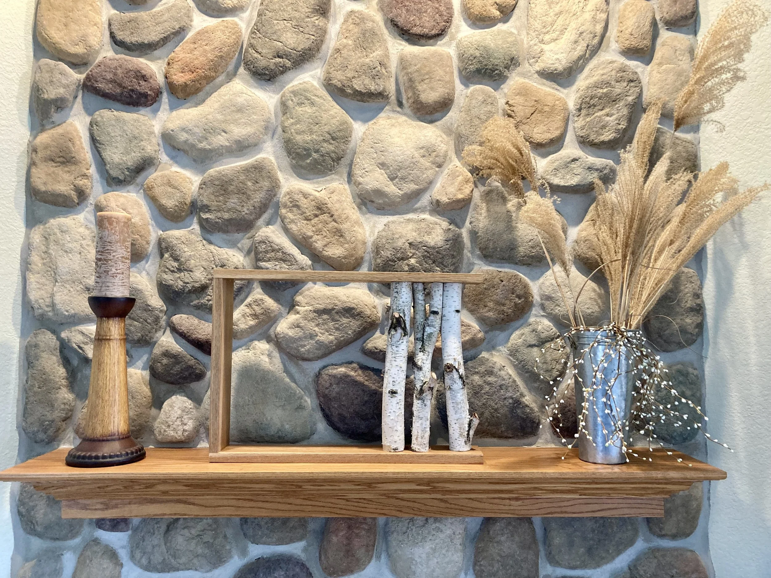 Decorative wooden shelf against a stone wall with a candle holder, a wooden frame, three bamboo sticks, and a silver vase with dried pampas grass and white berry branches.