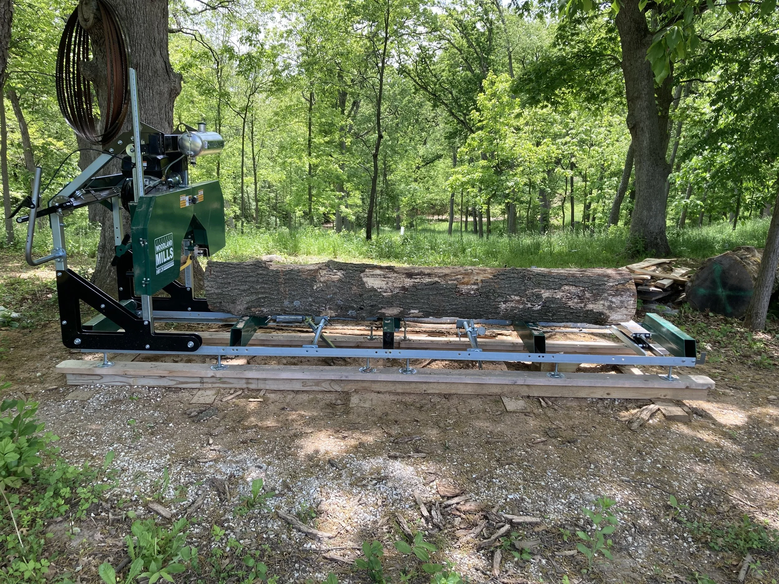 A mechanical sawmill cutting a log in a wooded outdoor area surrounded by trees.