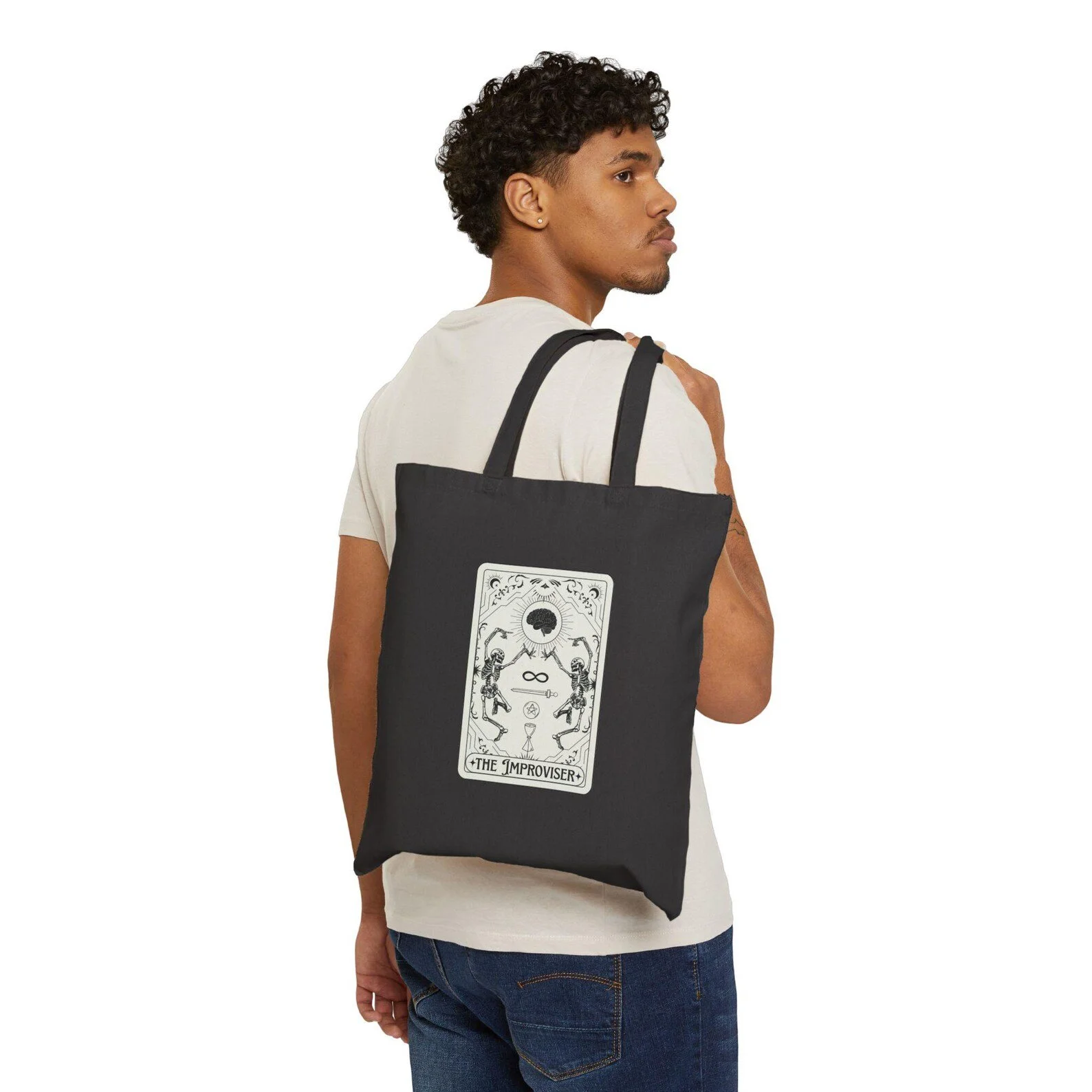A young man with curly hair and a mustache looking to the right, carrying a black tote bag with a tarot card design, wearing a white t-shirt and blue jeans.