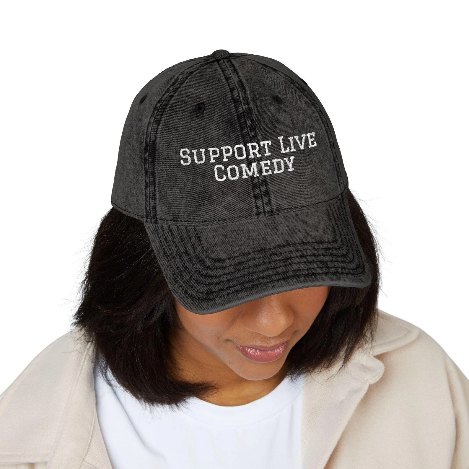 Person wearing a gray baseball cap with the words "Support Live Comedy" embroidered in white, with dark hair and a beige clothing.