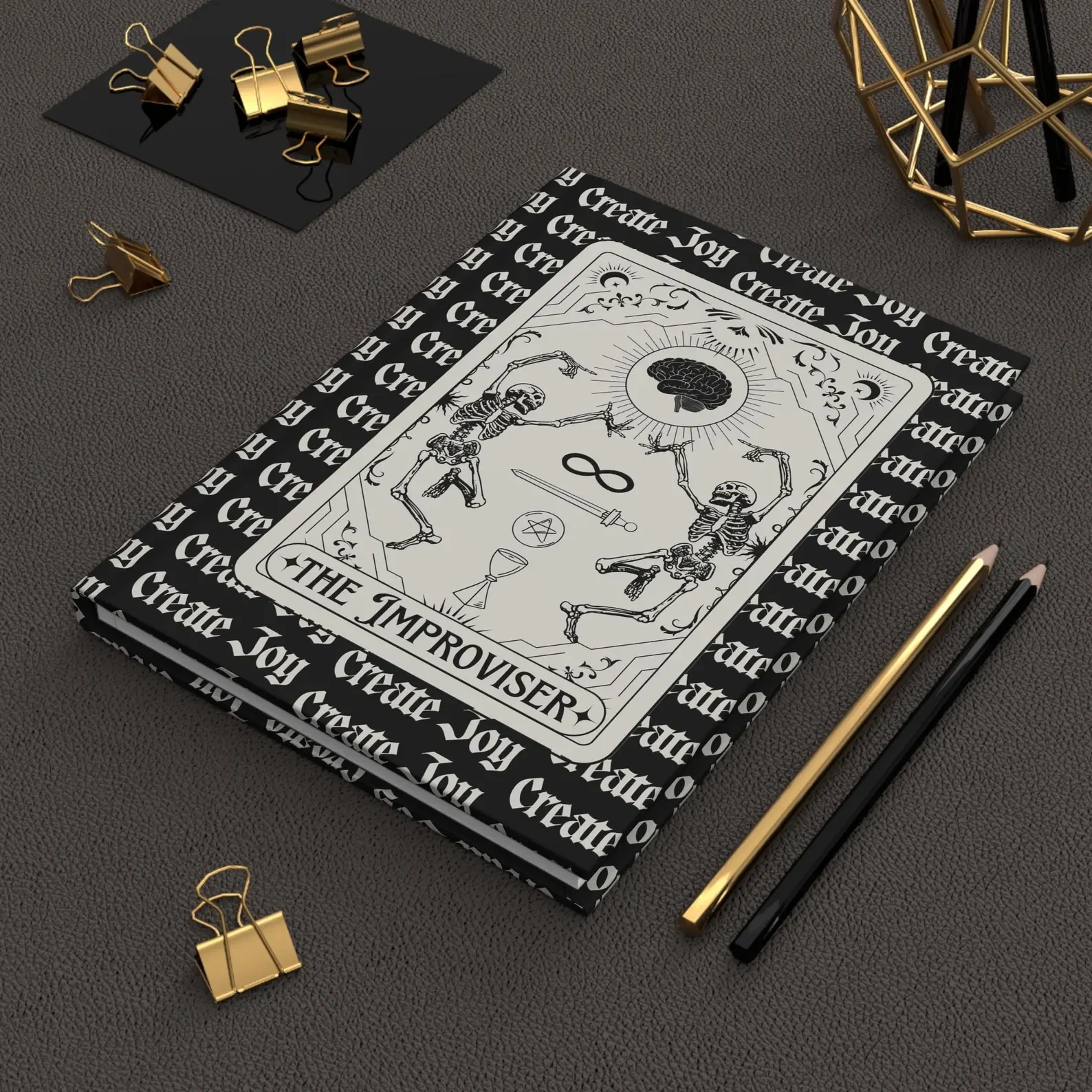A tarot card titled' placed on top of a book with a black and white cover that has decorative writing 'Create Joy' repeated around the border. The tarot card features illustrations of skeletons dancing under a sun with a brain at the center, a sword, a tea cup with a pentagram, and a snake.