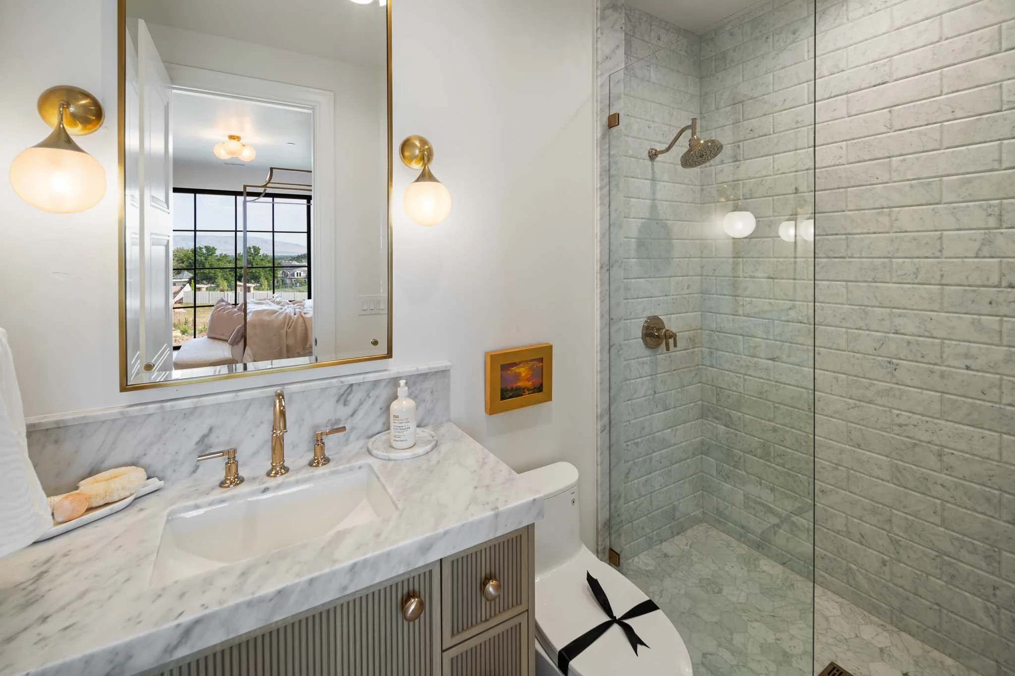 Modern bathroom with marble countertop, gold fixtures, wall-mounted light fixtures, large mirror, and a walk-in shower with white subway tile and a glass partition.