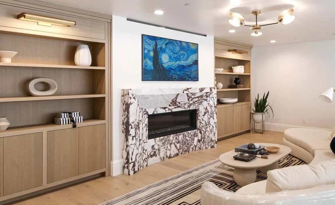 Living room with built-in wooden shelves, a marble fireplace, a painting above the fireplace, a modern ceiling light fixture, a large white sectional sofa, a round coffee table, and a potted plant.