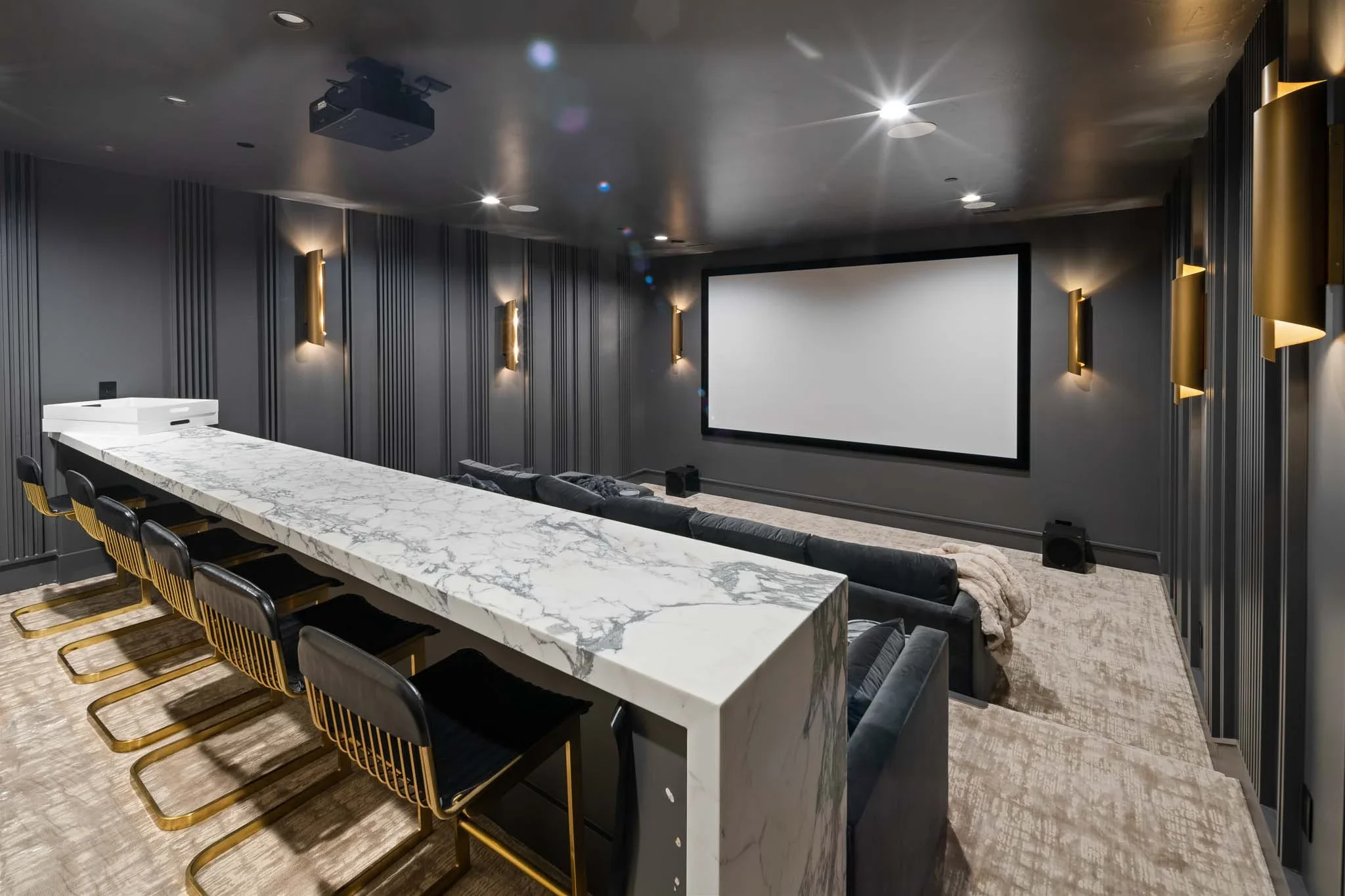 A modern home theater room with a large white marble-top table, black chairs with gold legs, a comfortable sofa with a blanket, wall-mounted gold lights, and a large screen on the wall.