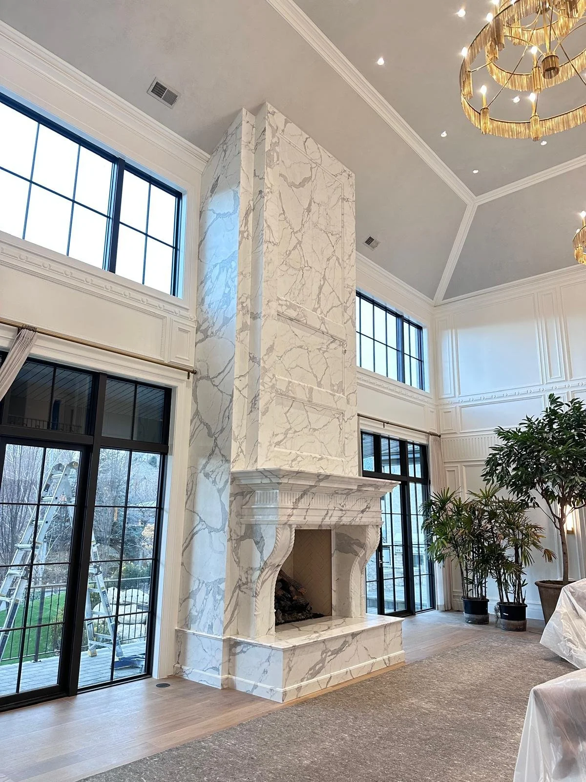 Interior of a luxury living room with a custom tall marble fireplace and large windows, decorated with plants, and featuring a gold chandelier hanging from the ceiling.