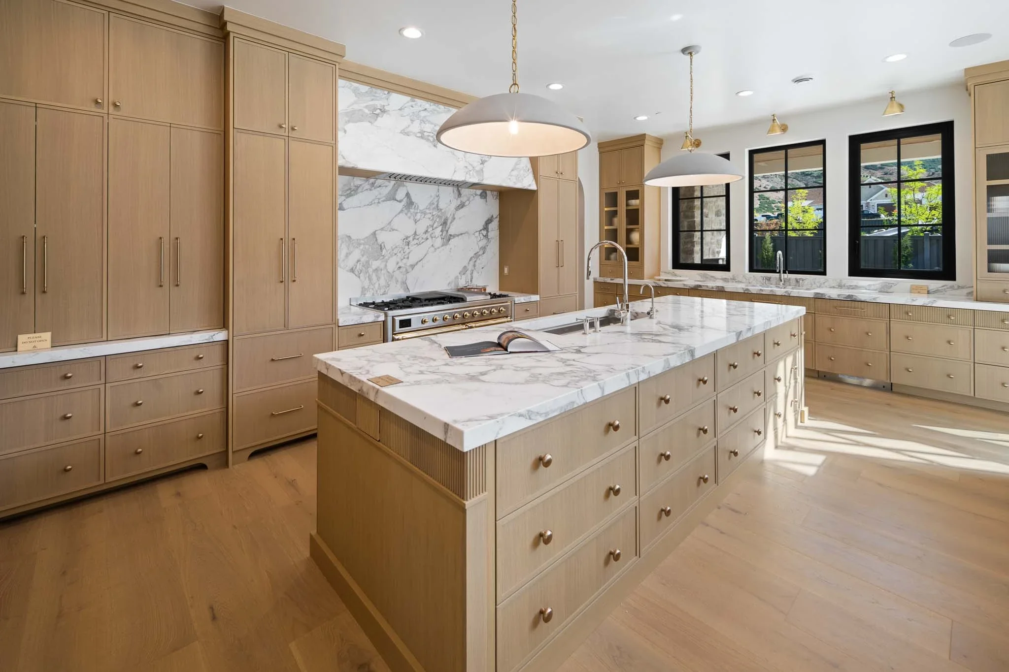A modern kitchen with light wood cabinets, a large island with a marble countertop, and a marble backsplash behind the stove. There are three large windows with black frames, and two pendant lights hanging over the island. The kitchen has lots of nat