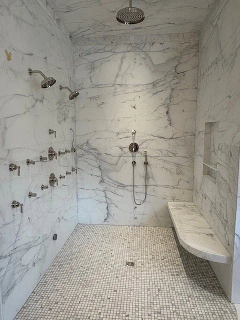 A custom luxury marble shower with white marble walls, multiple showerheads on the left wall, and a built-in marble bench on the right.