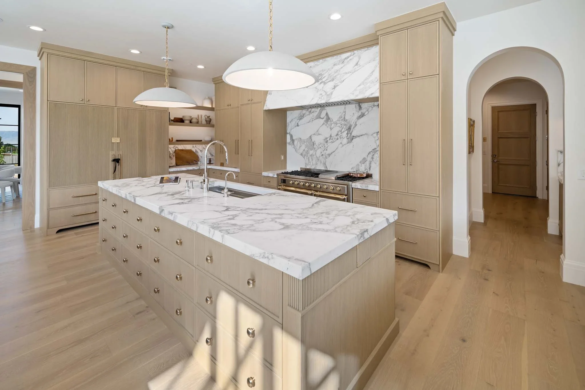 Modern kitchen with beige cabinetry, marble countertops, and a marble backsplash, featuring stainless steel stove and two large white pendant lights hanging over the island.