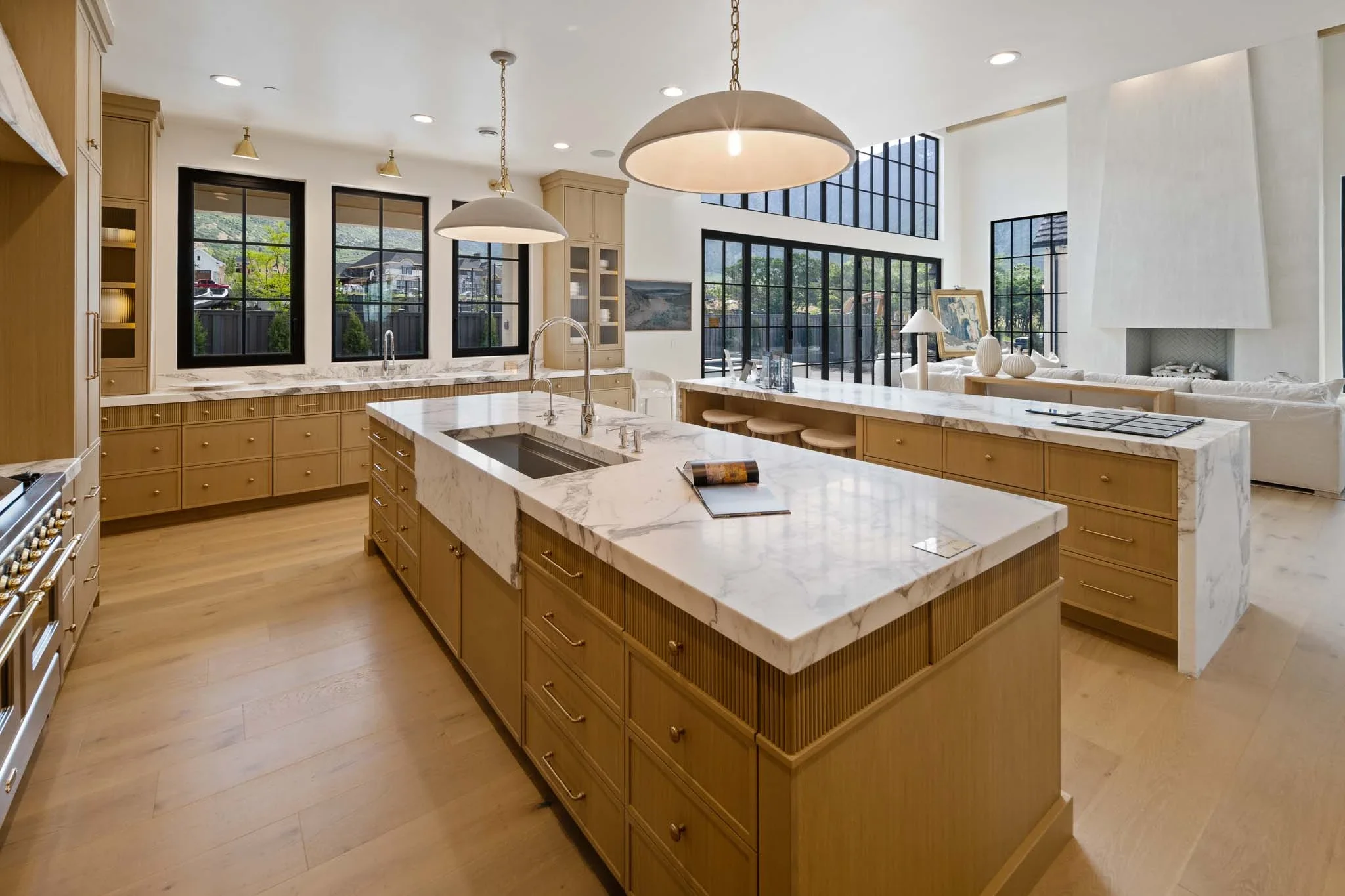 Modern kitchen with large marble island, light wood cabinets, black-framed windows, and a living room with a fireplace and large windows in the background.