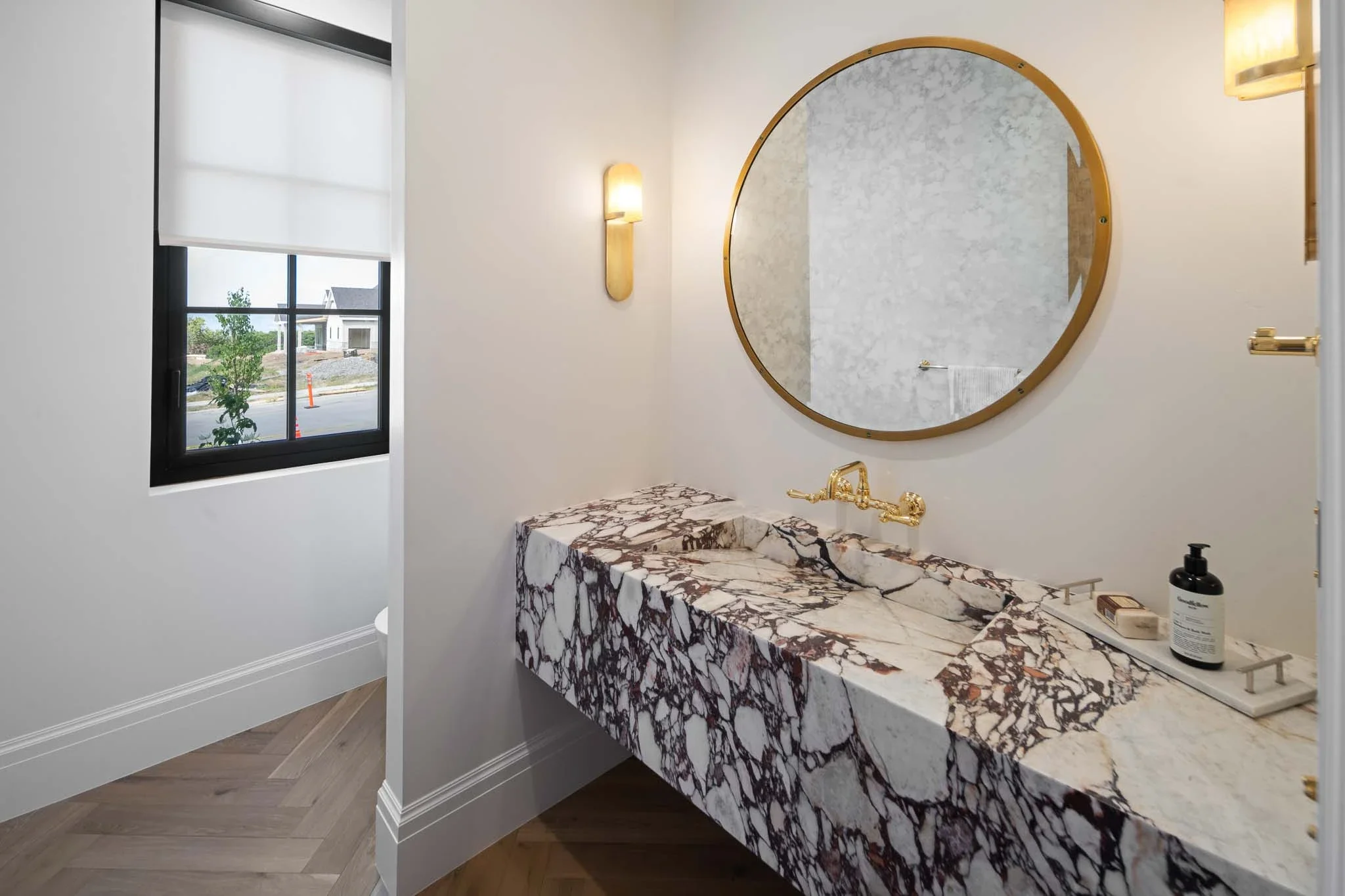 A bathroom with a marble vanity, a large round mirror with a gold frame, and a wall sconce light. There is a black-framed window with a white roller shade, and a small outside view of some trees and buildings. On the vanity, there is a bottle of hand
