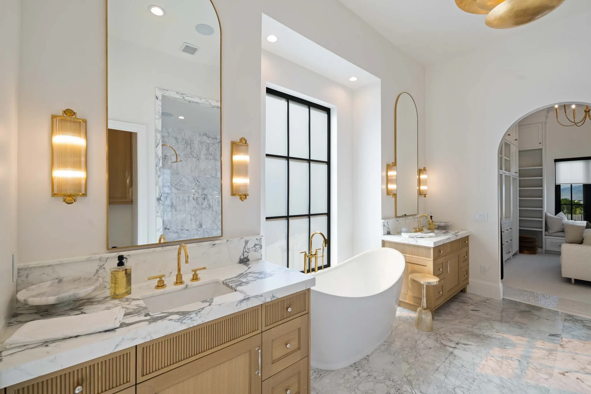 Modern bathroom with white walls, marble countertops, a freestanding bathtub, gold fixtures, large mirrors, and a large window with black framing.