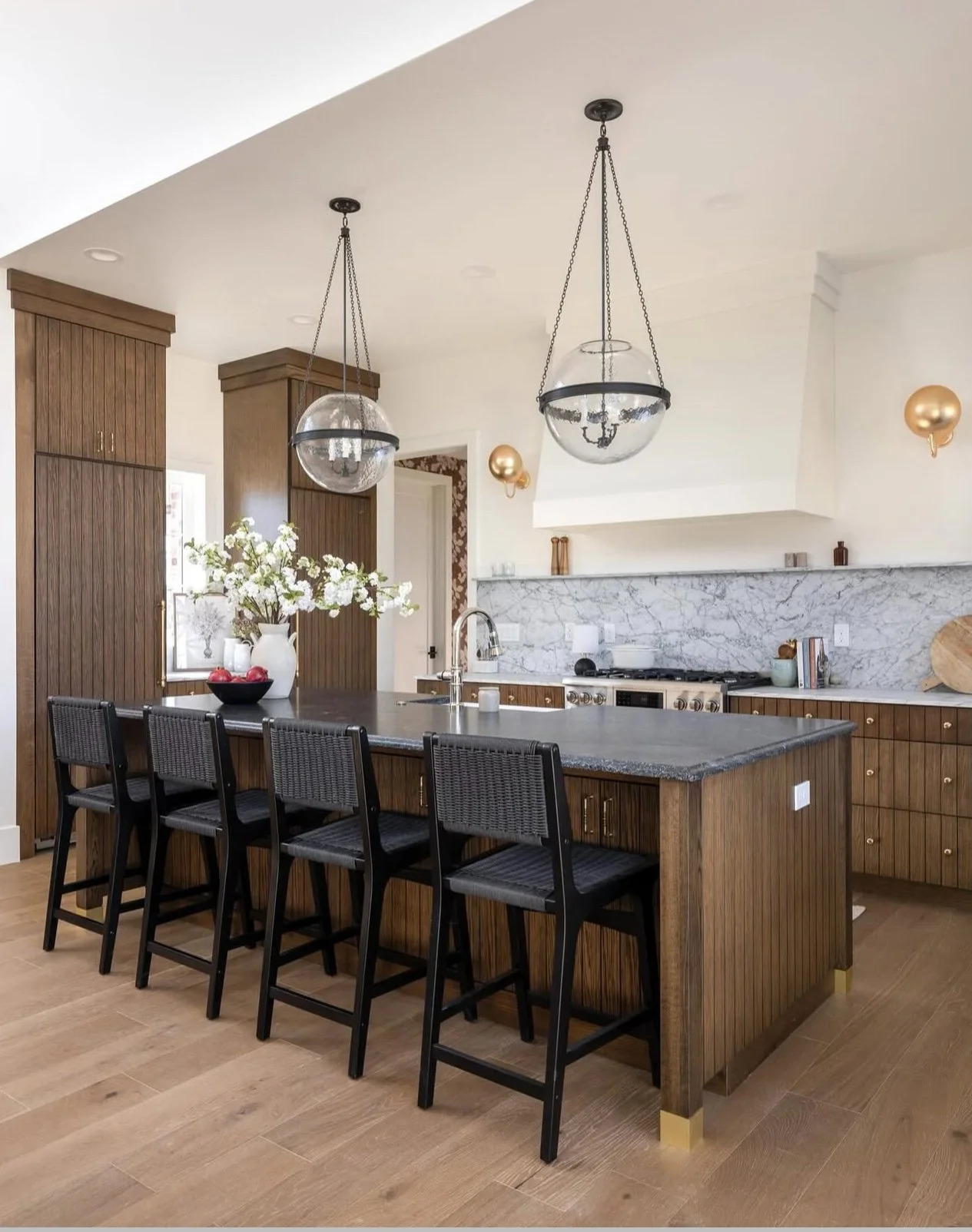 Modern kitchen with wooden cabinets, marble backsplash, black bar stools at a dark countertop island, and glass pendant lights hanging from the ceiling.