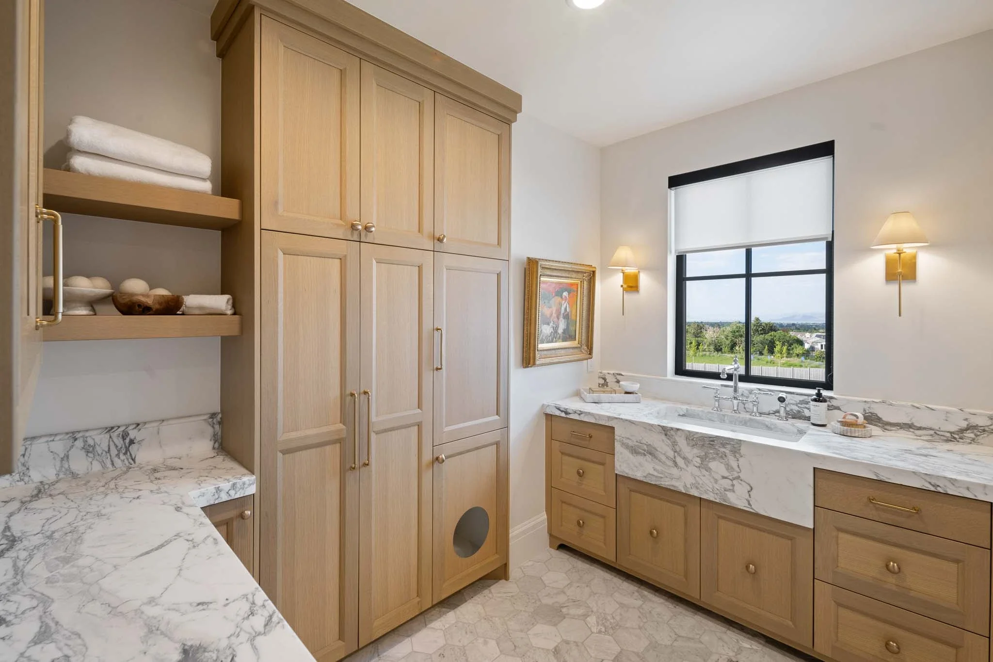 A bathroom with light wood cabinets, a white marble countertop, and a window showing a view of trees and houses outside. There are wall-mounted lights, a framed painting, and folded towels on a wooden shelf.