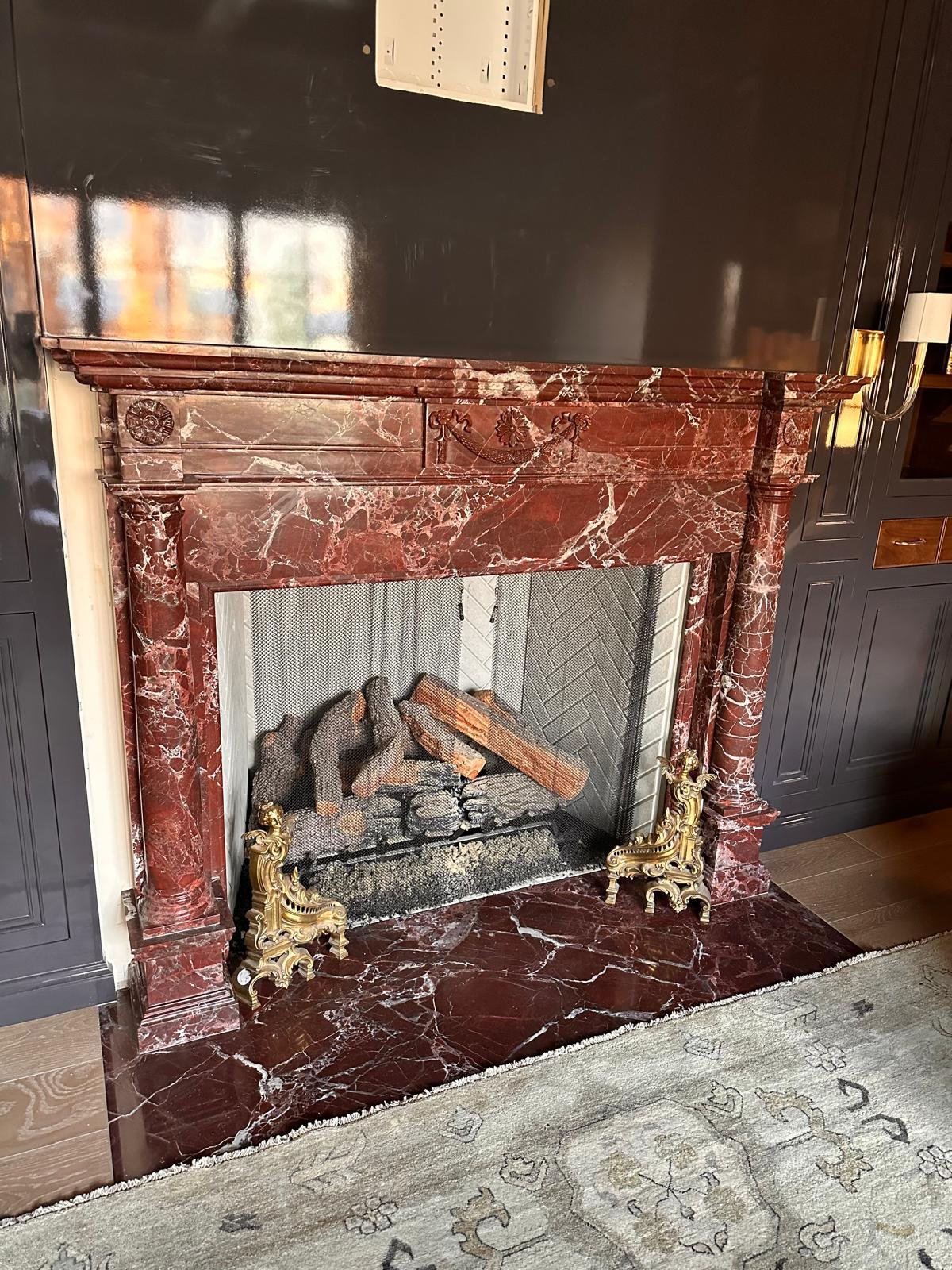 A red custom marble fireplace with decorative gold accents, set against a dark wall and a light-colored rug with a pattern.