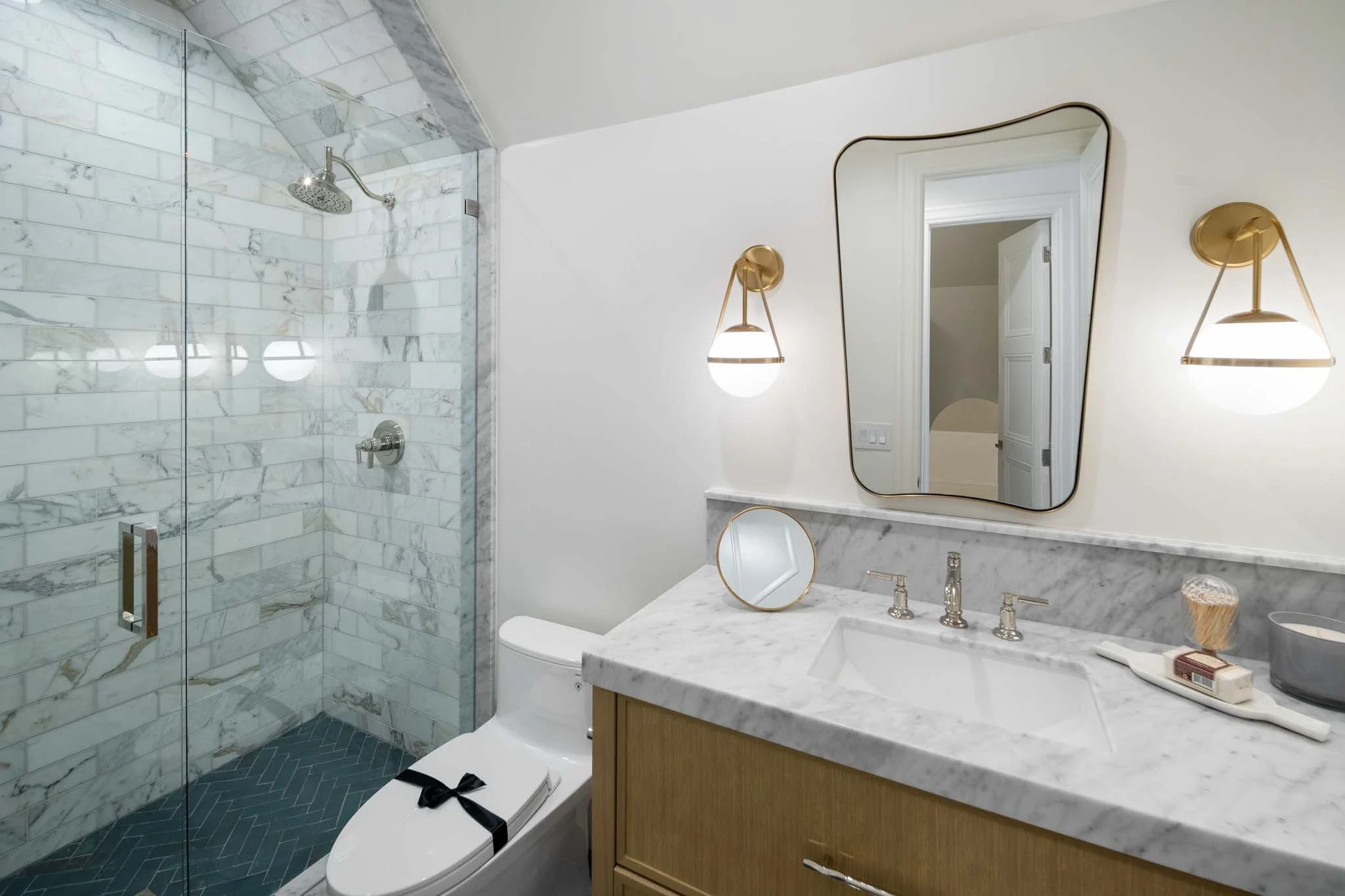 Modern bathroom with marble counters, a large mirror, decorative brass sconces, a glass shower with marble tiling, and a toilet with a ribbon on it.