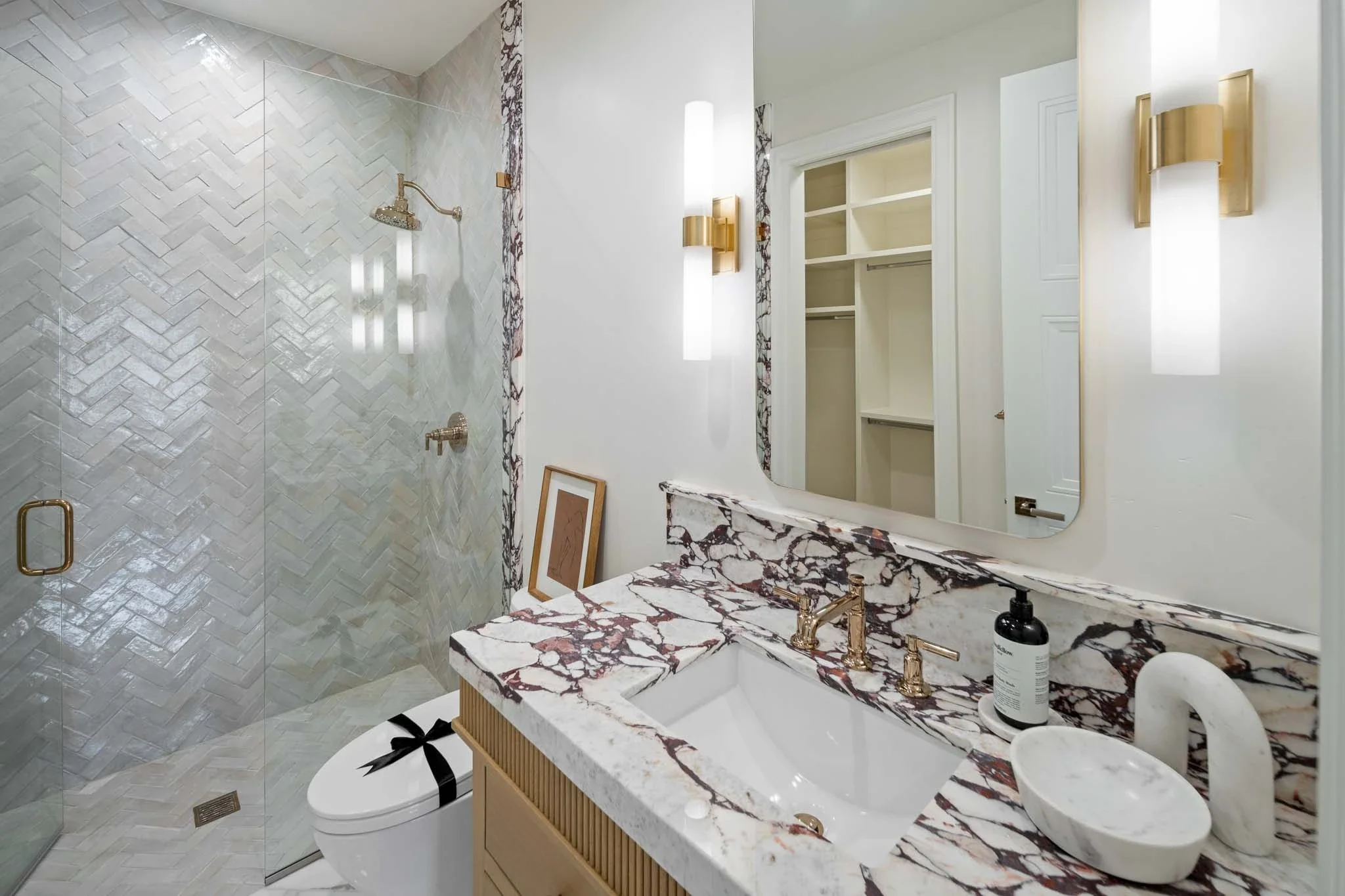 Modern bathroom with marble sink countertop, gold fixtures, large mirror, and walk-in shower with herringbone tiled wall.
