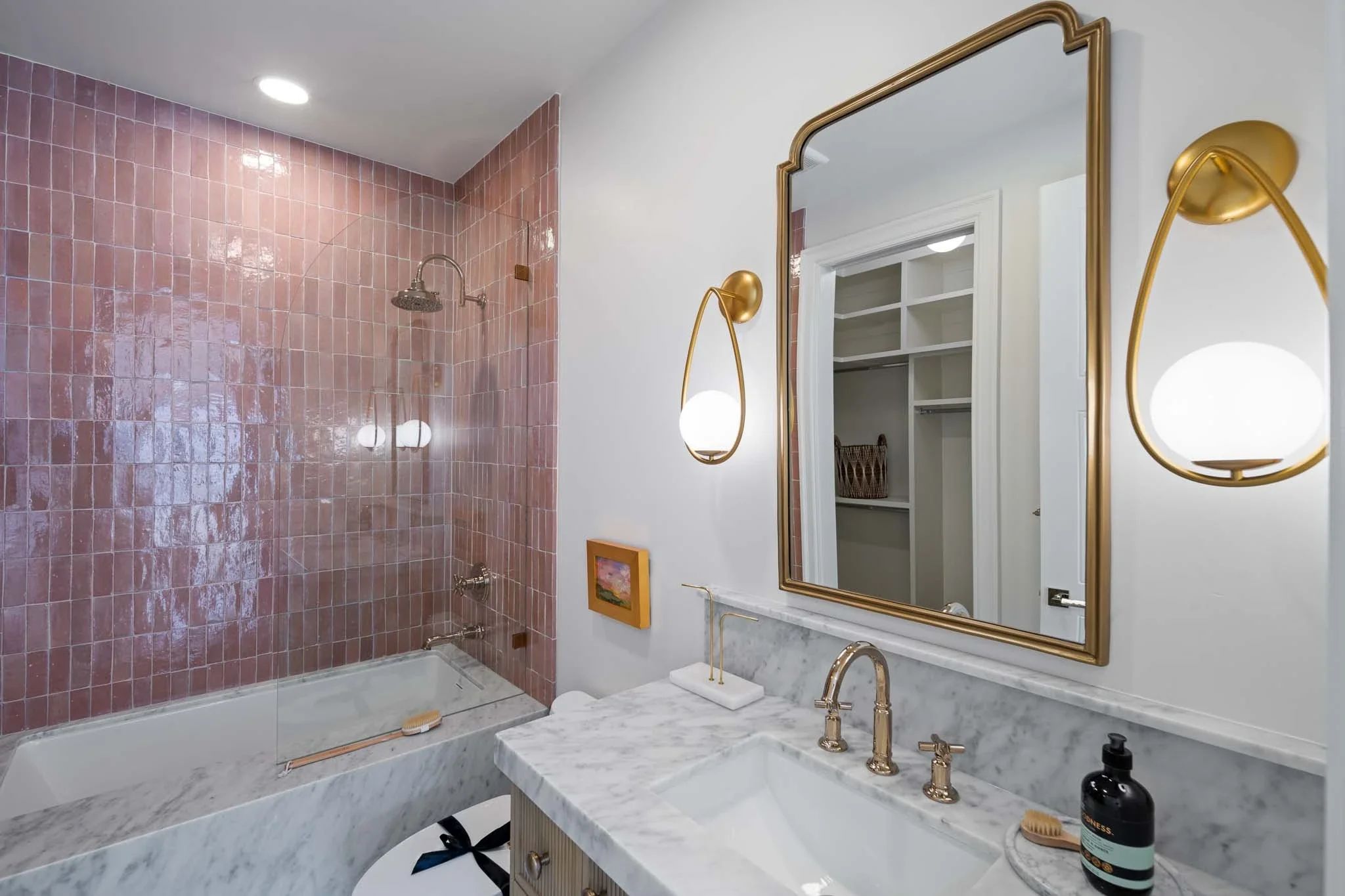Bathroom with pink tiled shower area, white walls, large mirror with gold trim, marble countertop with gold fixtures, and a open closet in the background.