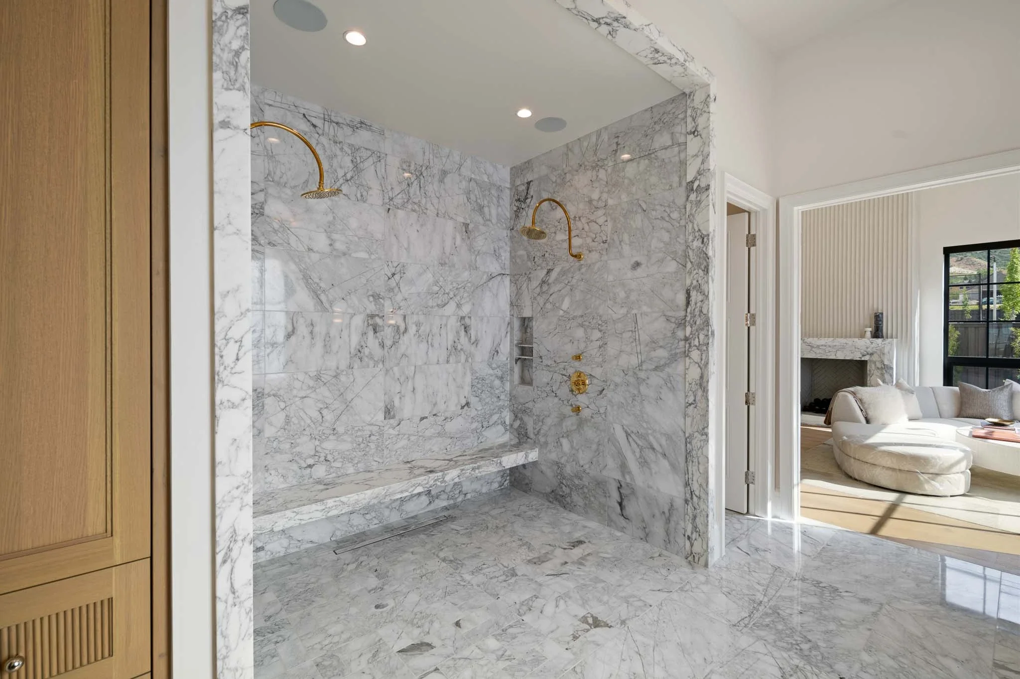 A modern bathroom with marble tiles on the floor, walls, and shower area. The shower has two gold fixtures with handheld heads. To the right, an open door reveals part of a living room with a white sofa and fireplace, and a window showing trees outsi