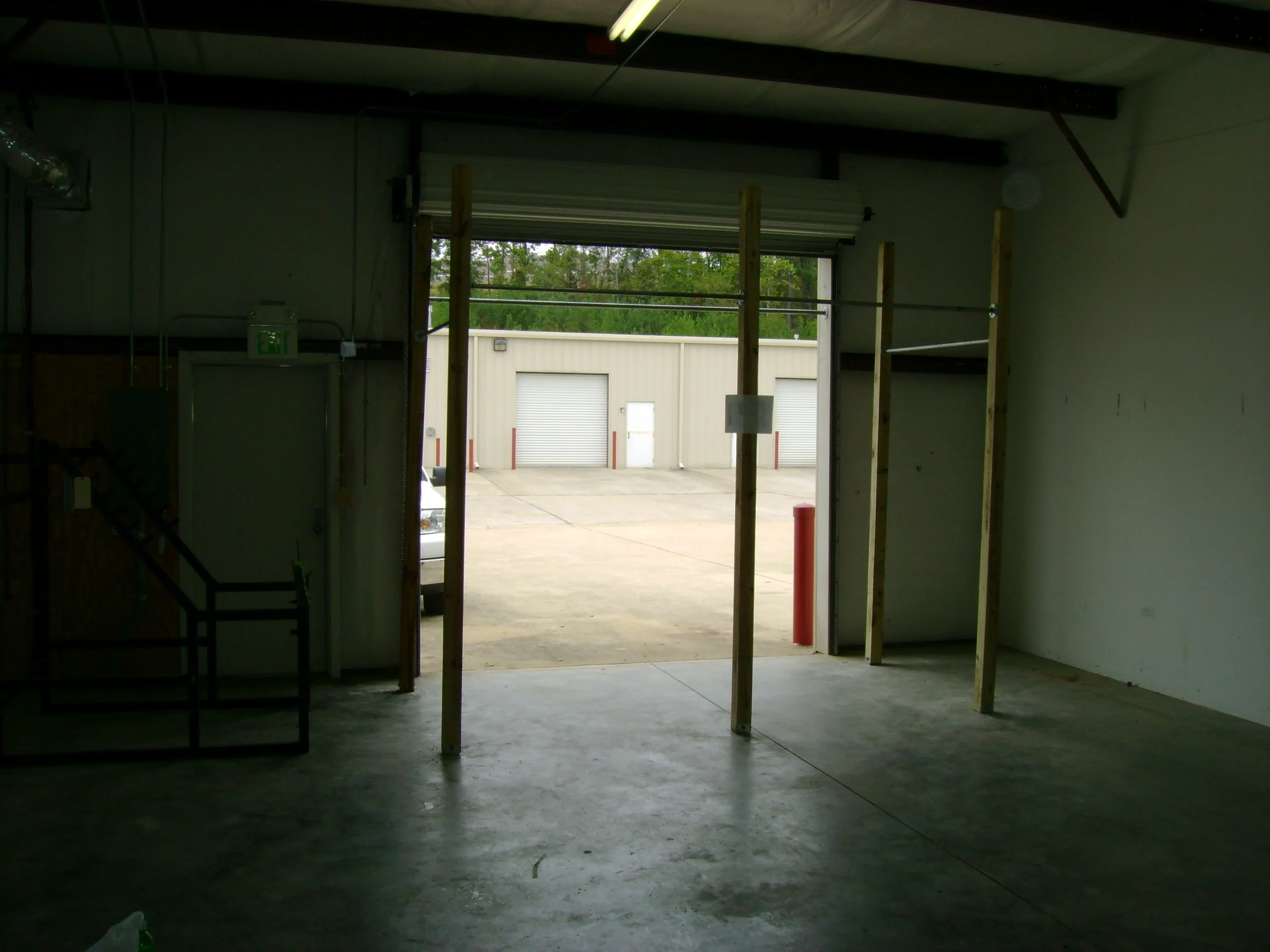 Interior view of a garage or workshop with unfinished walls and ceiling, showing wooden studs and an open garage door leading outside, revealing a paved area and a building with closed garage doors.