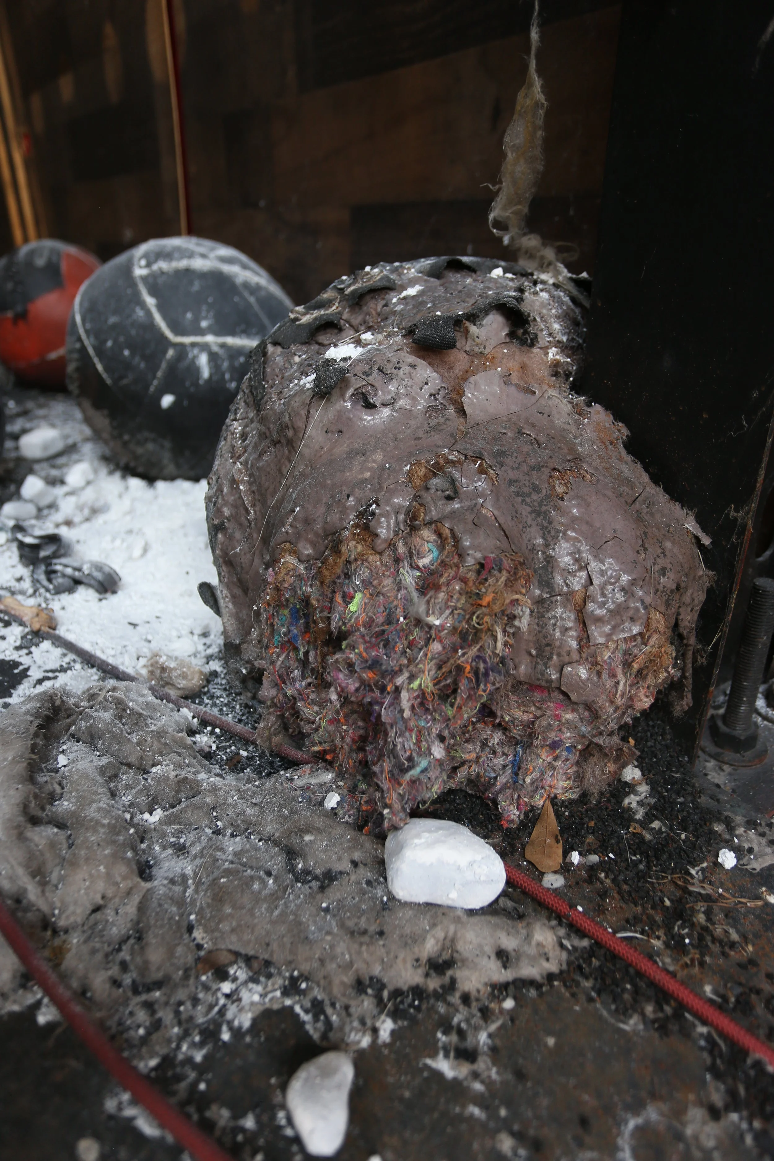 Burnt remnants of a large object, with melted and charred materials, ash, and debris scattered around, including a white piece of ice or foam, on a dark surface with some equipment and wood paneling in the background.