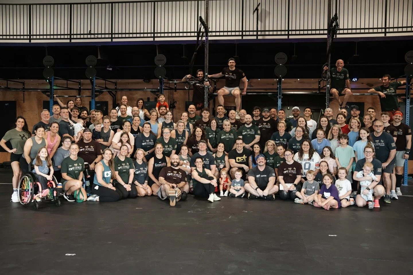Group photo of diverse people at a CrossFit gym, including children, adults, and seniors, with some on workout equipment or sitting on the floor, smiling and posing for the camera.