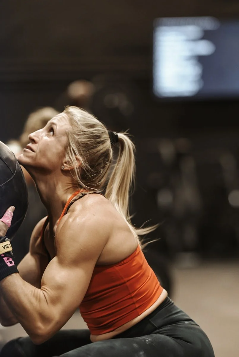 A woman with blonde hair in a ponytail, wearing a red tank top, is lifting a black weight in a gym.