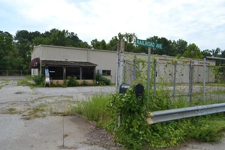 An outdoor scene featuring an old building behind a chain-link fence, with a person holding an umbrella, and a street sign for Railroad Ave.