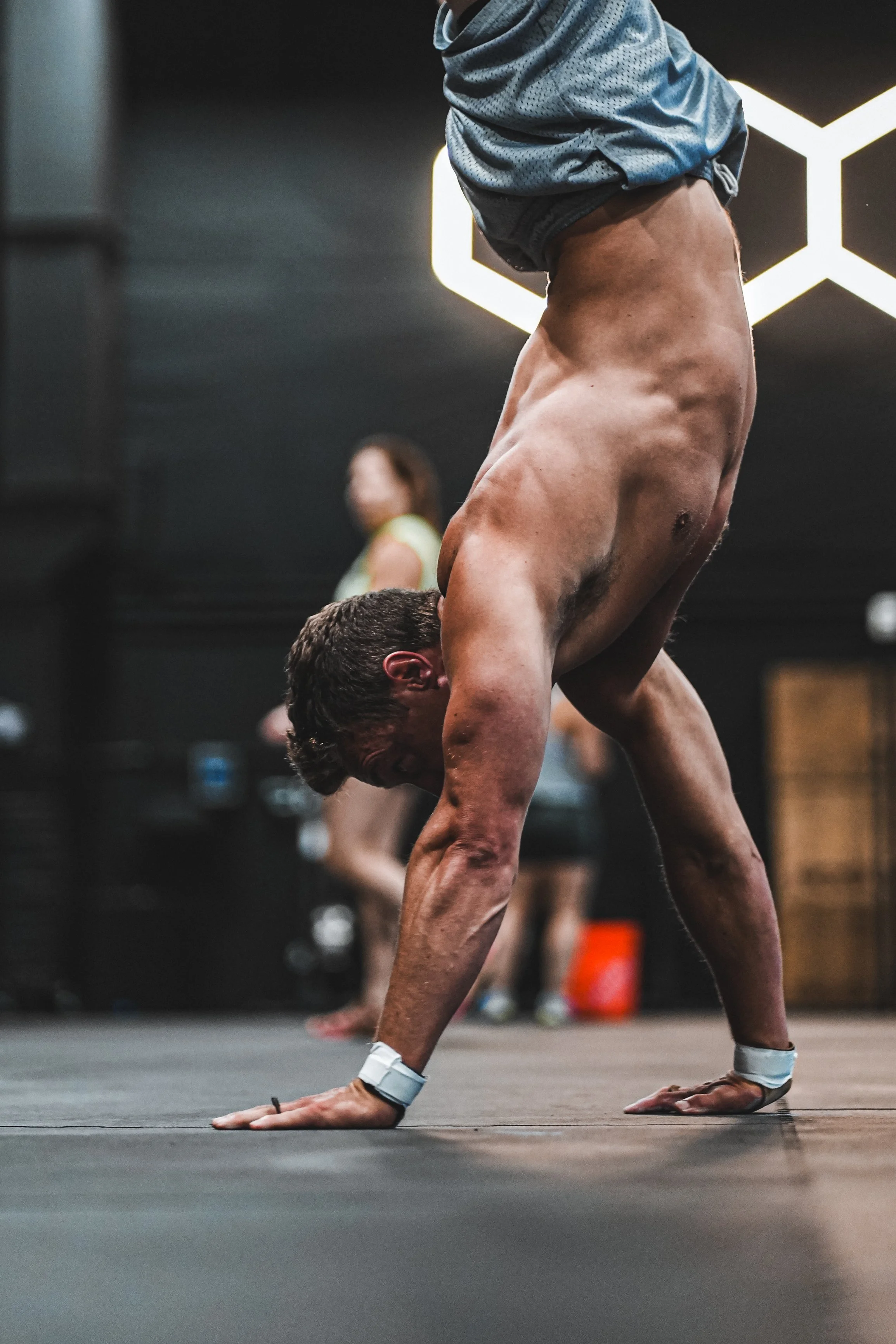 A man performing a plank exercise on his hands and feet in a gym, with a woman in the background and illuminated geometric wall art.