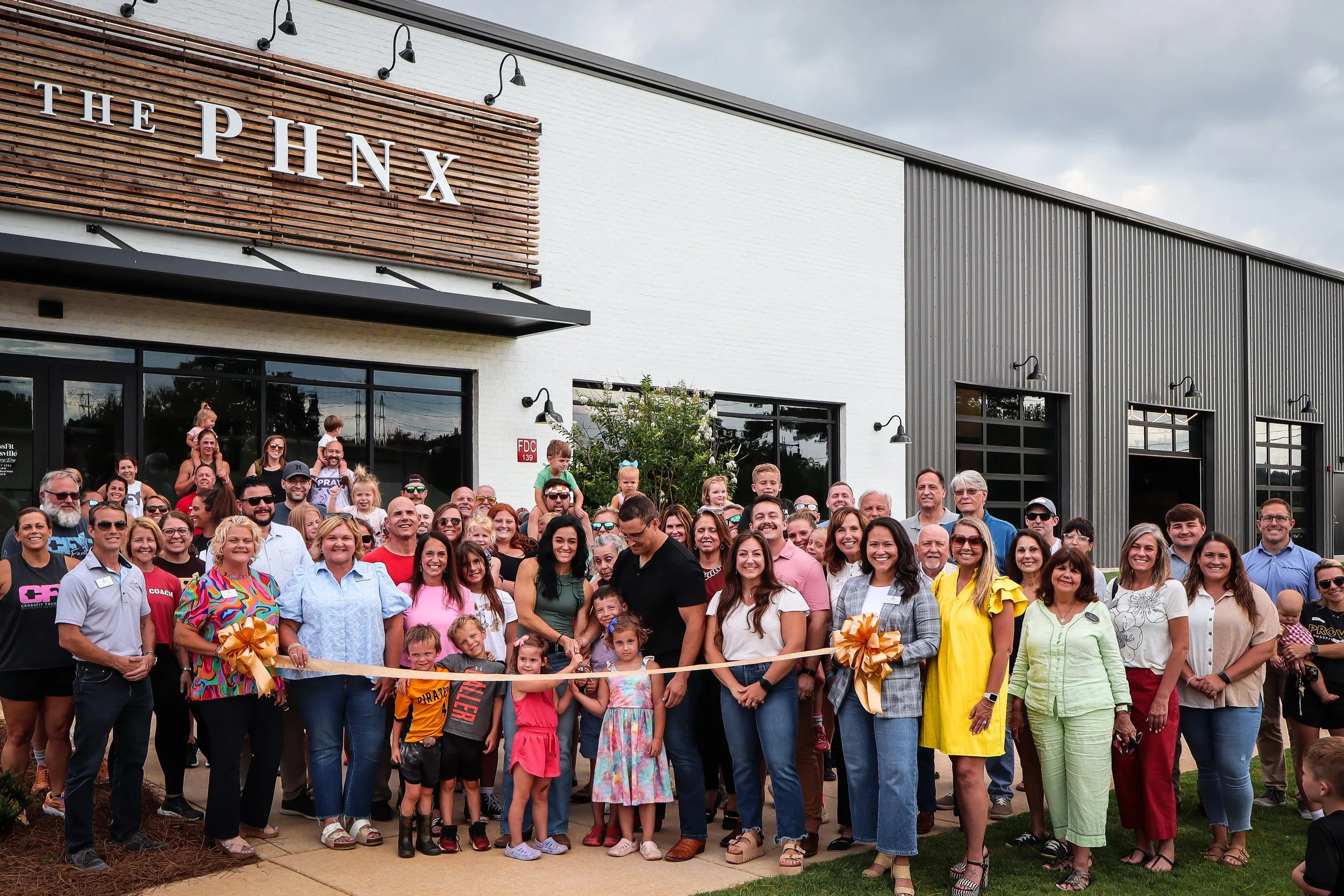 A large group of diverse people gathered in front of a building with a sign that reads 'THE PHNX'. The group appears to be celebrating, and some are holding a ribbon with bows, indicating a ribbon-cutting event. The building has modern industrial-style architecture with white and gray exterior walls and large windows.