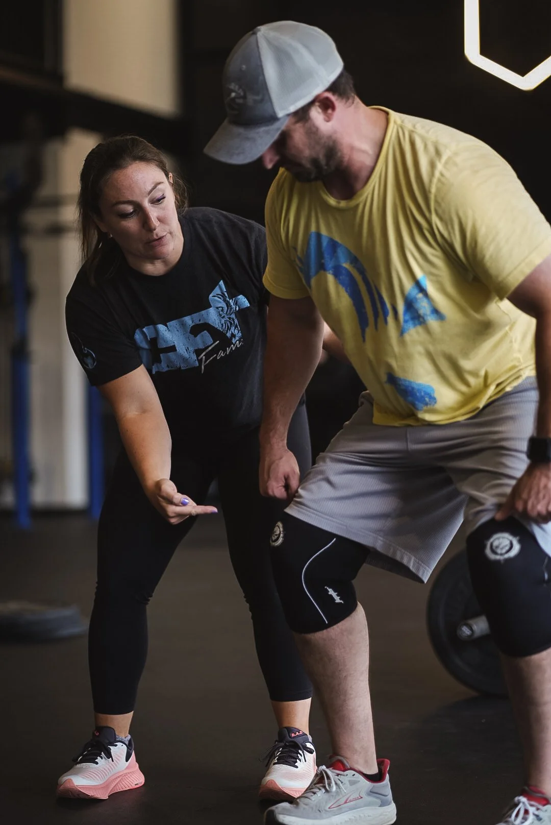 Two people, a woman and a man, are in a gym, engaged in a coaching or training session. The woman is pointing and explaining something, and they are both leaning towards each other, focused on the activity. The woman is wearing a black T-shirt with a