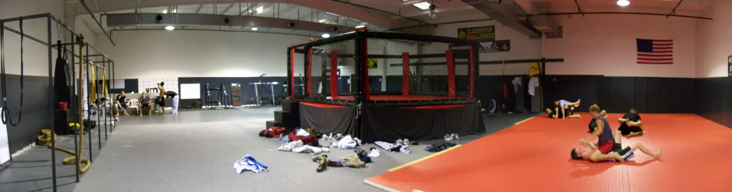 Inside a martial arts training gym with a red mat area, a cage for fighting, some people practicing grappling, and clothing scattered on the floor.