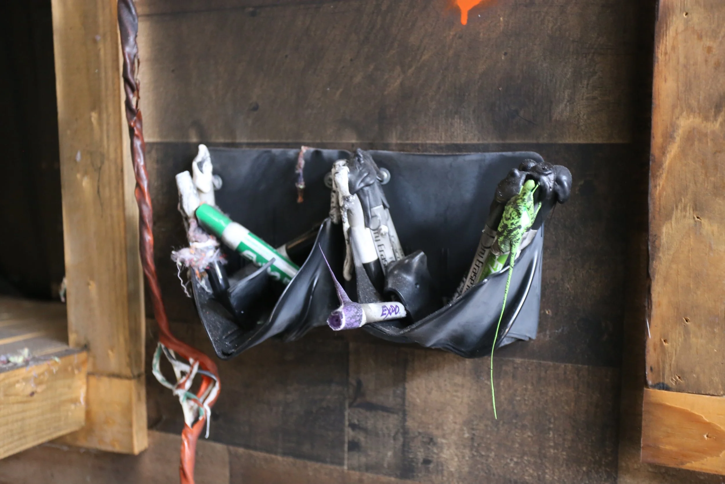 View of a black toolbox containing various tools, including a green-handled marker, a black and white clamp, and a purple handle tool, placed inside a wooden storage area with visible wood framing and side panel.