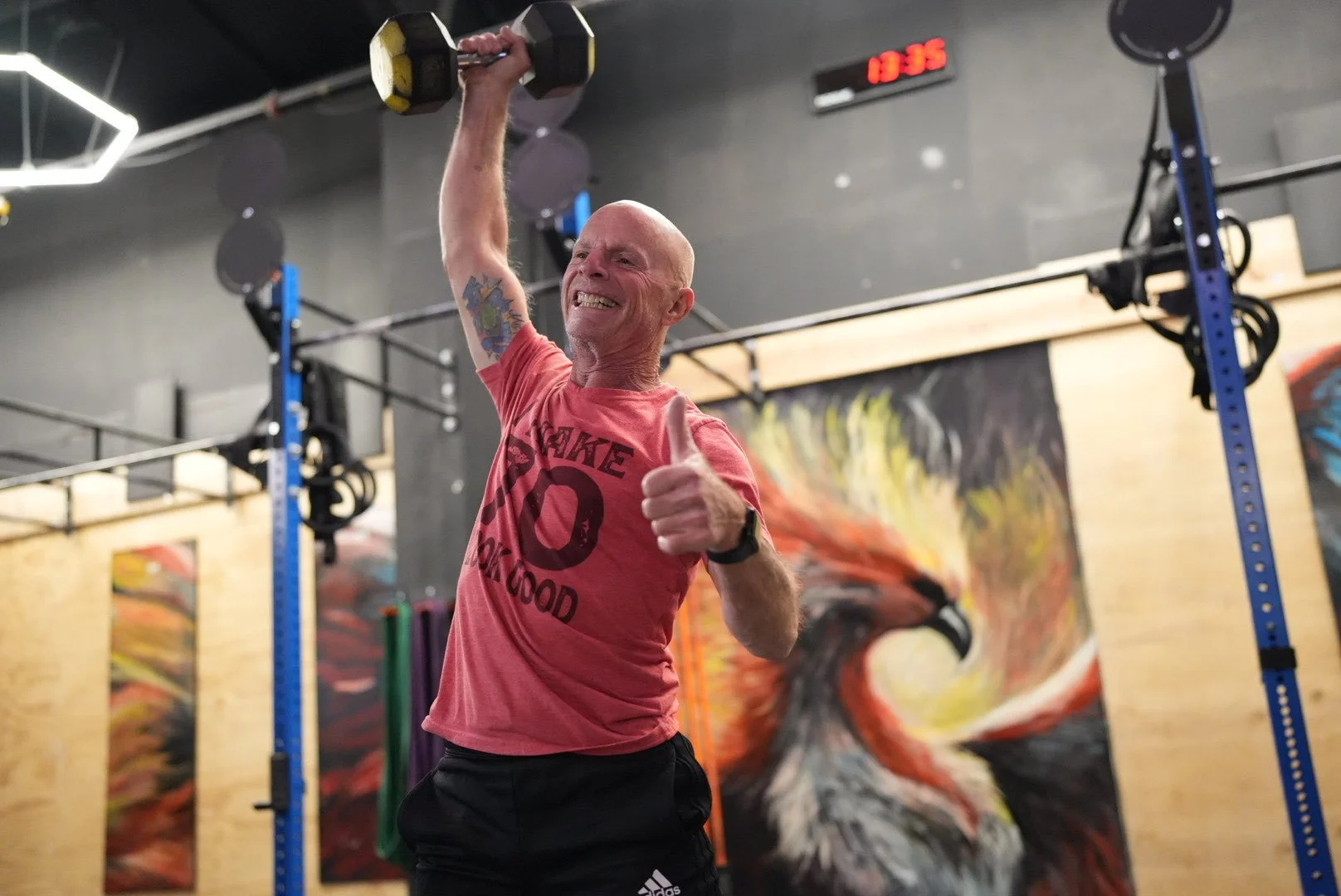 A man with a shaved head and tattoos, wearing a red t-shirt that says 'Shake O' and 'Bakwood,' lifting a dumbbell in a gym. He is giving a thumbs up and smiling. The gym has black walls, a large colorful artwork of a horse, and a digital clock showing the time 13:35.
