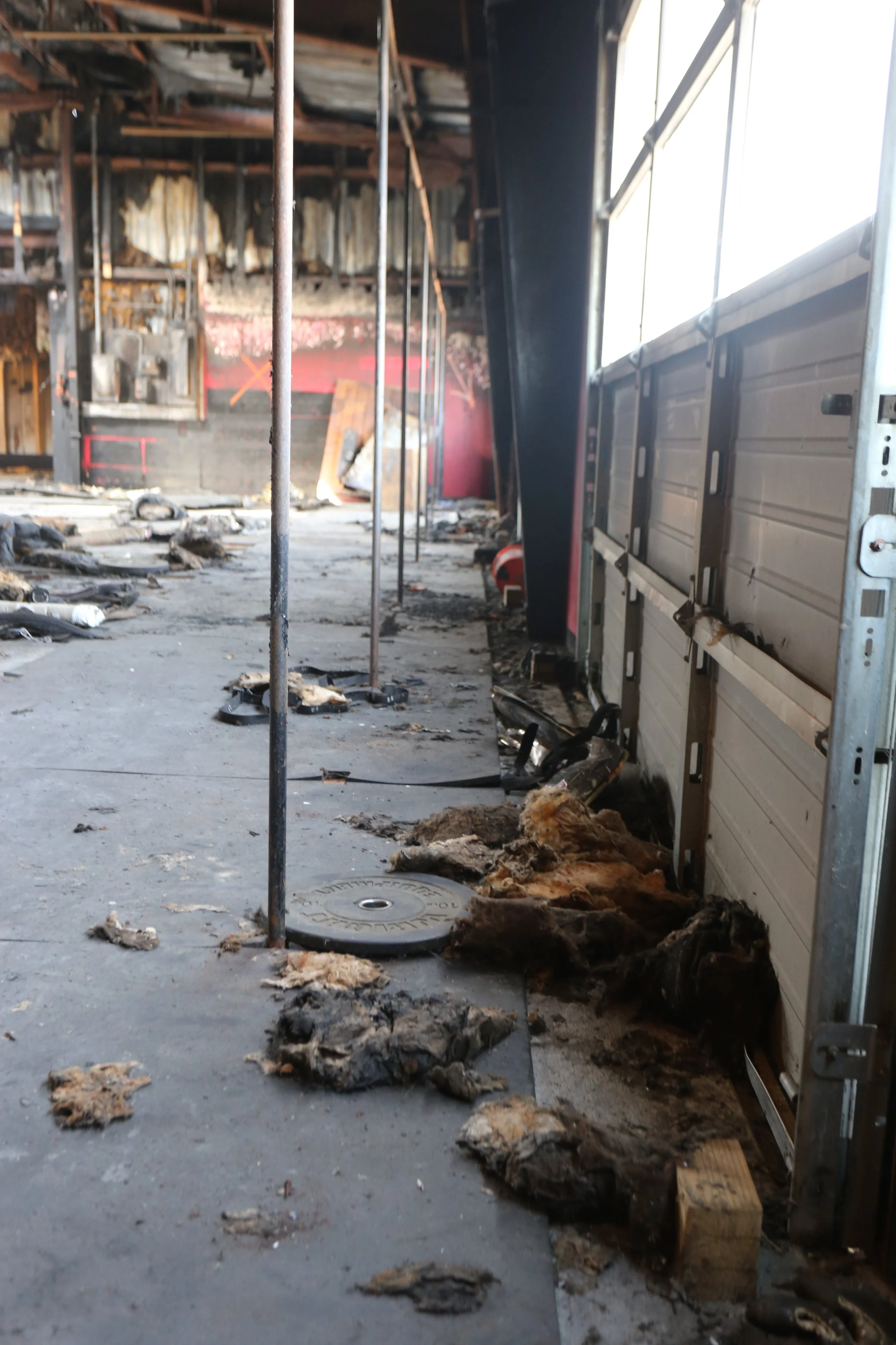 An interior view of a building that has experienced fire damage. The space is filled with debris, soot, and damaged insulation. Metal support poles are visible, and the walls and ceiling show extensive burn marks and charring. The garage door on the 