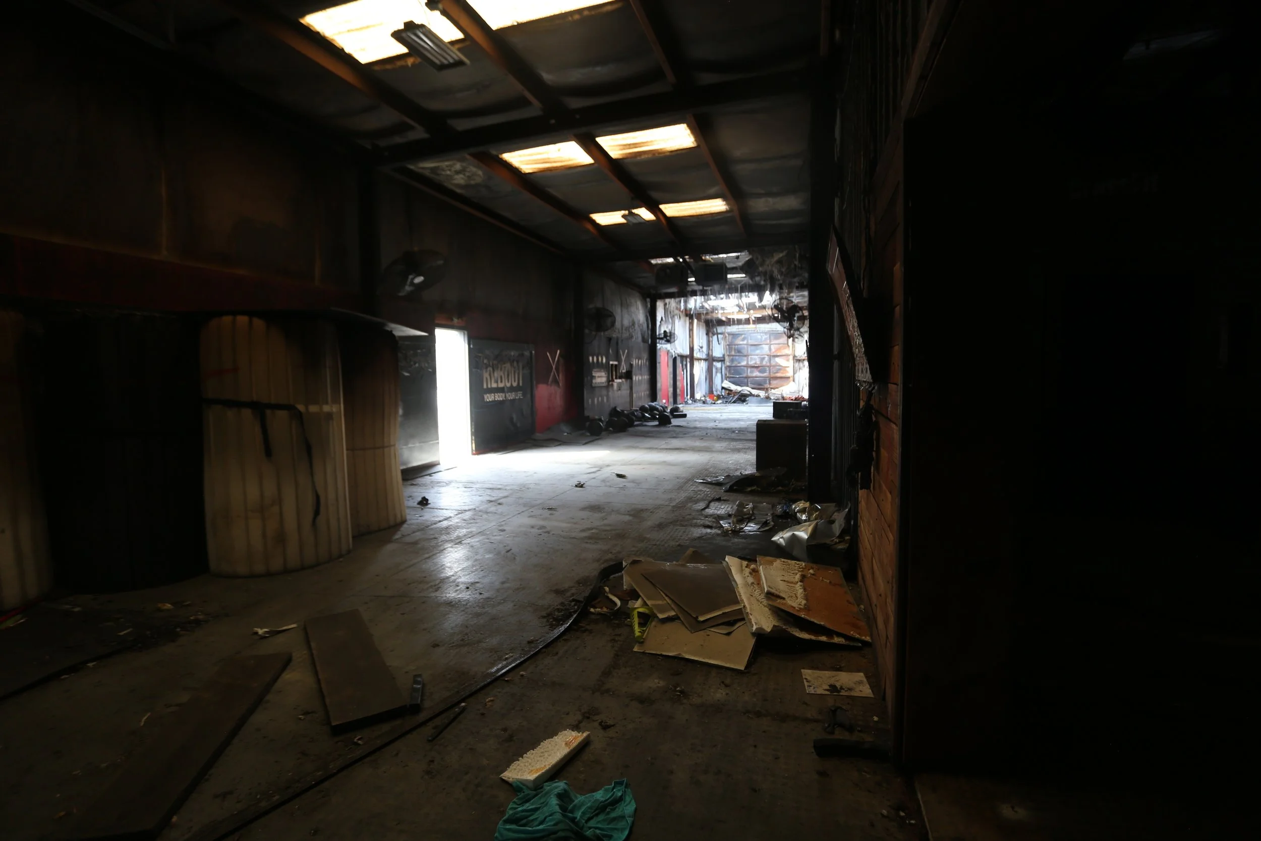 The interior of a damaged, abandoned building with a dark, charred appearance, debris scattered across the floor, and sunlight shining through broken roof panels.
