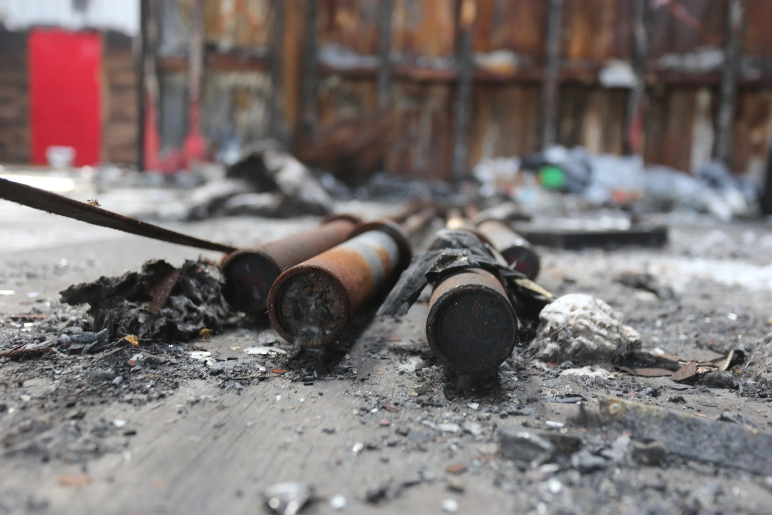 Close-up of burnt and rusted metal pipes on the ground with ashes and debris, blurred background of a damaged wooden fence with snow patches.