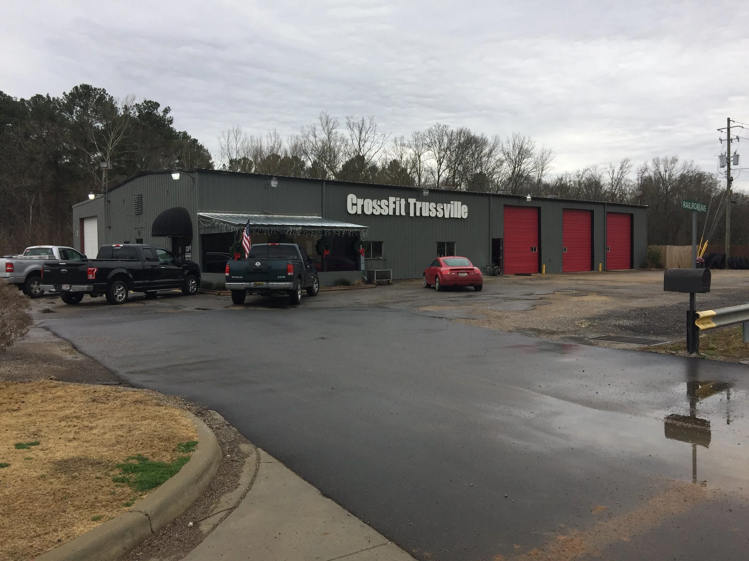 Exterior of a building with a sign that says 'CrossFit Trussville,' parked cars, a flag with Christmas decorations, and a parking lot with puddles.