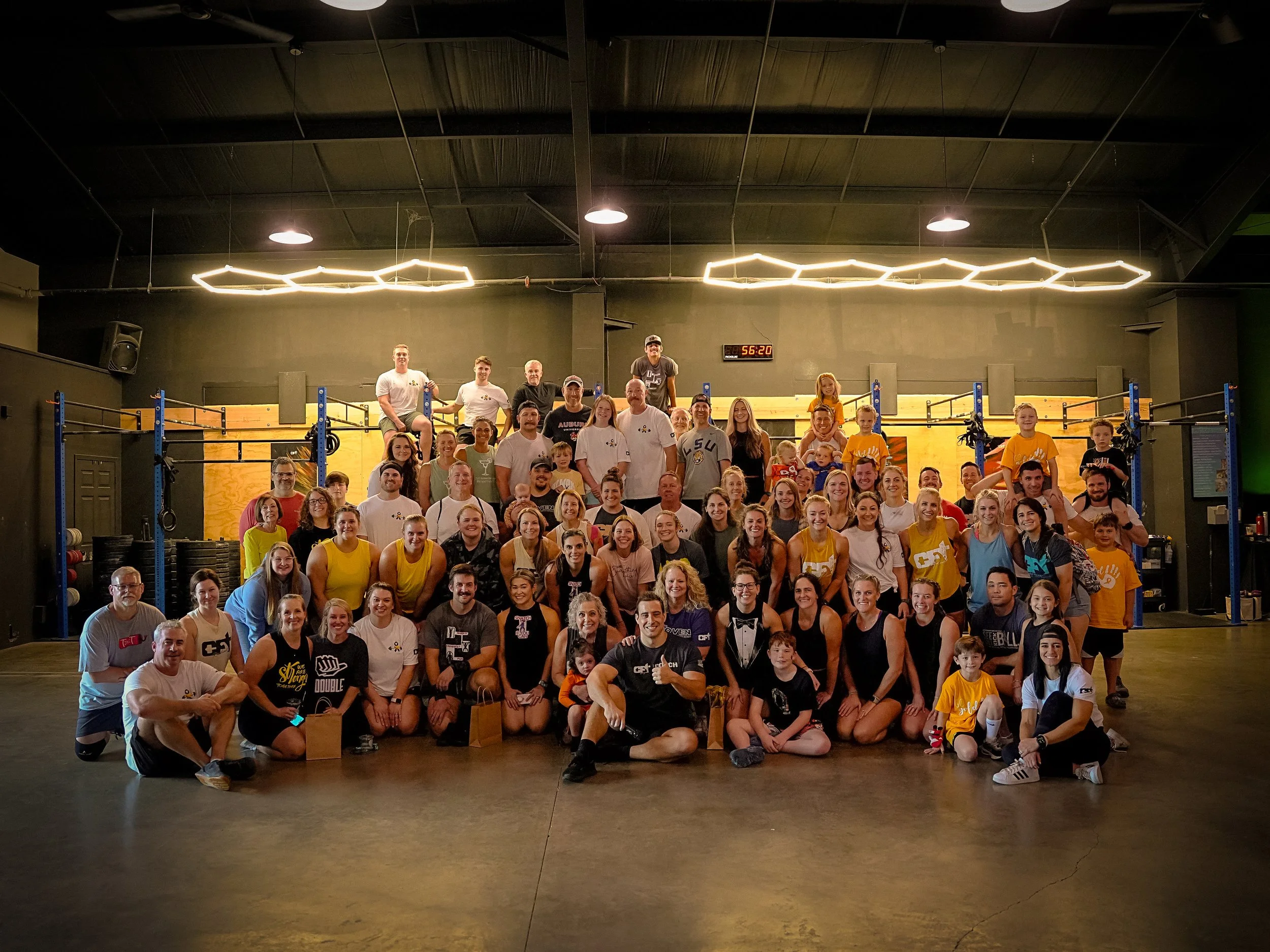 Large group photo of diverse people in workout clothes inside a gym, some sitting, others standing, smiling at the camera, with gym equipment and workout rigs in the background.