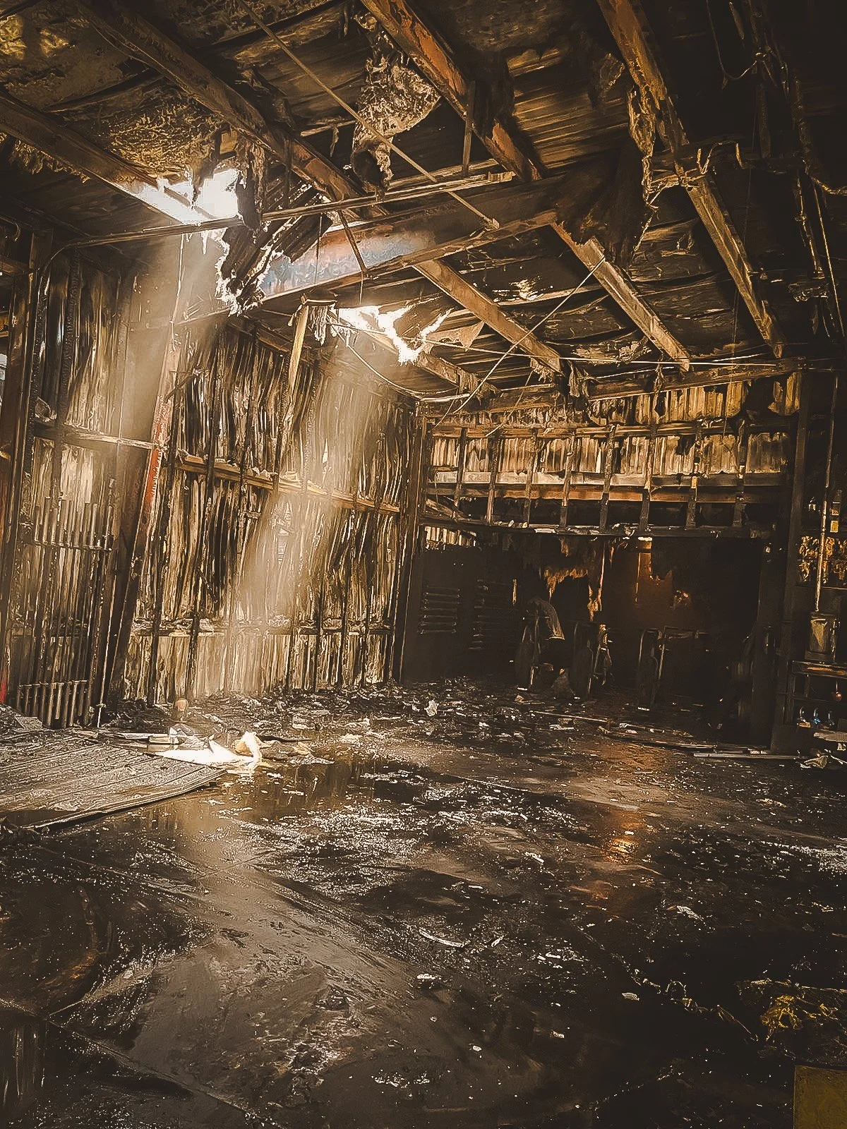 A room with burned and charred walls and ceiling, showing significant fire damage with blackened debris on the floor and burnt materials hanging from the ceiling.
