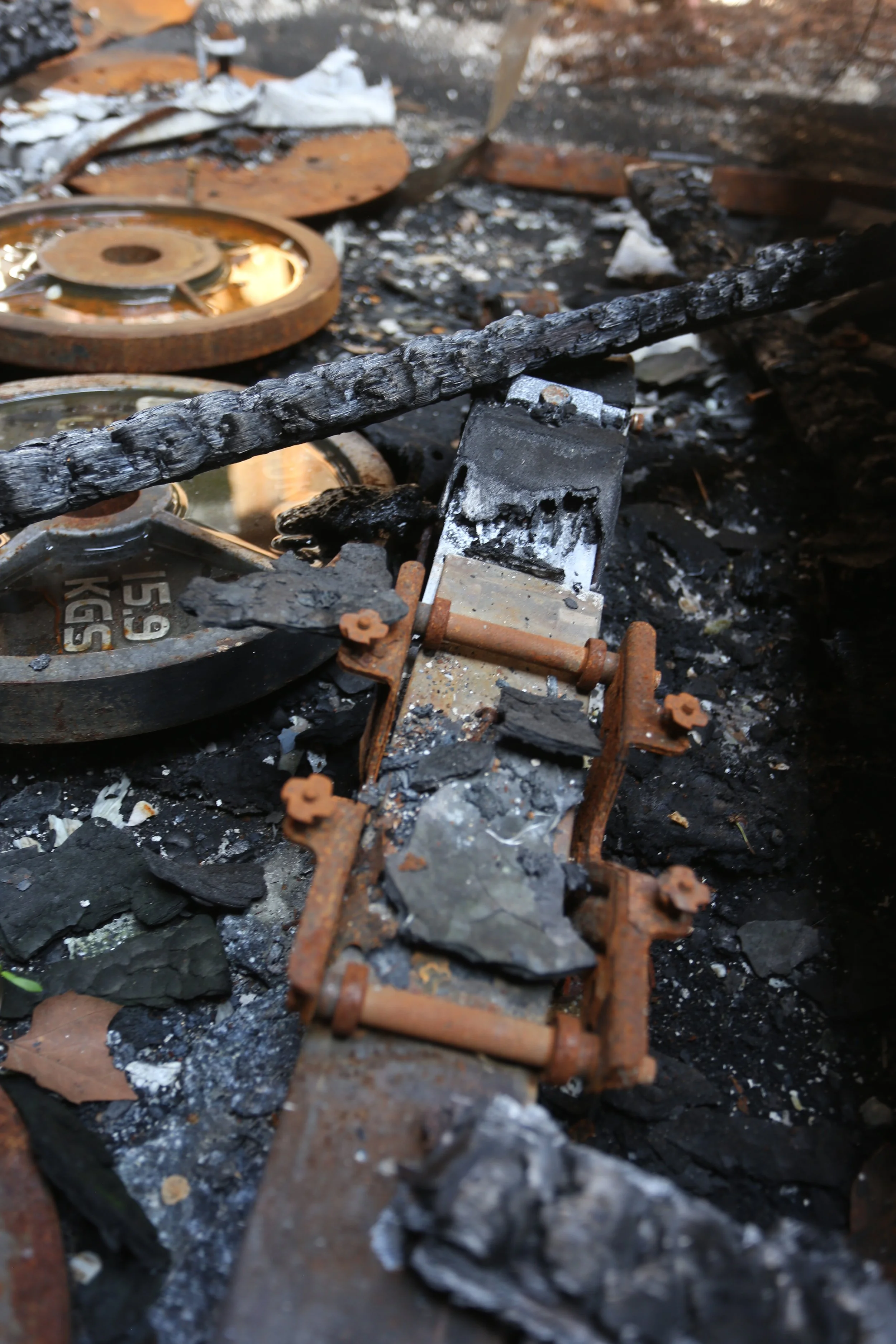Burned and charred remains of a mechanical or electronic device, surrounded by ashes and debris, indicating a recent fire or destruction.