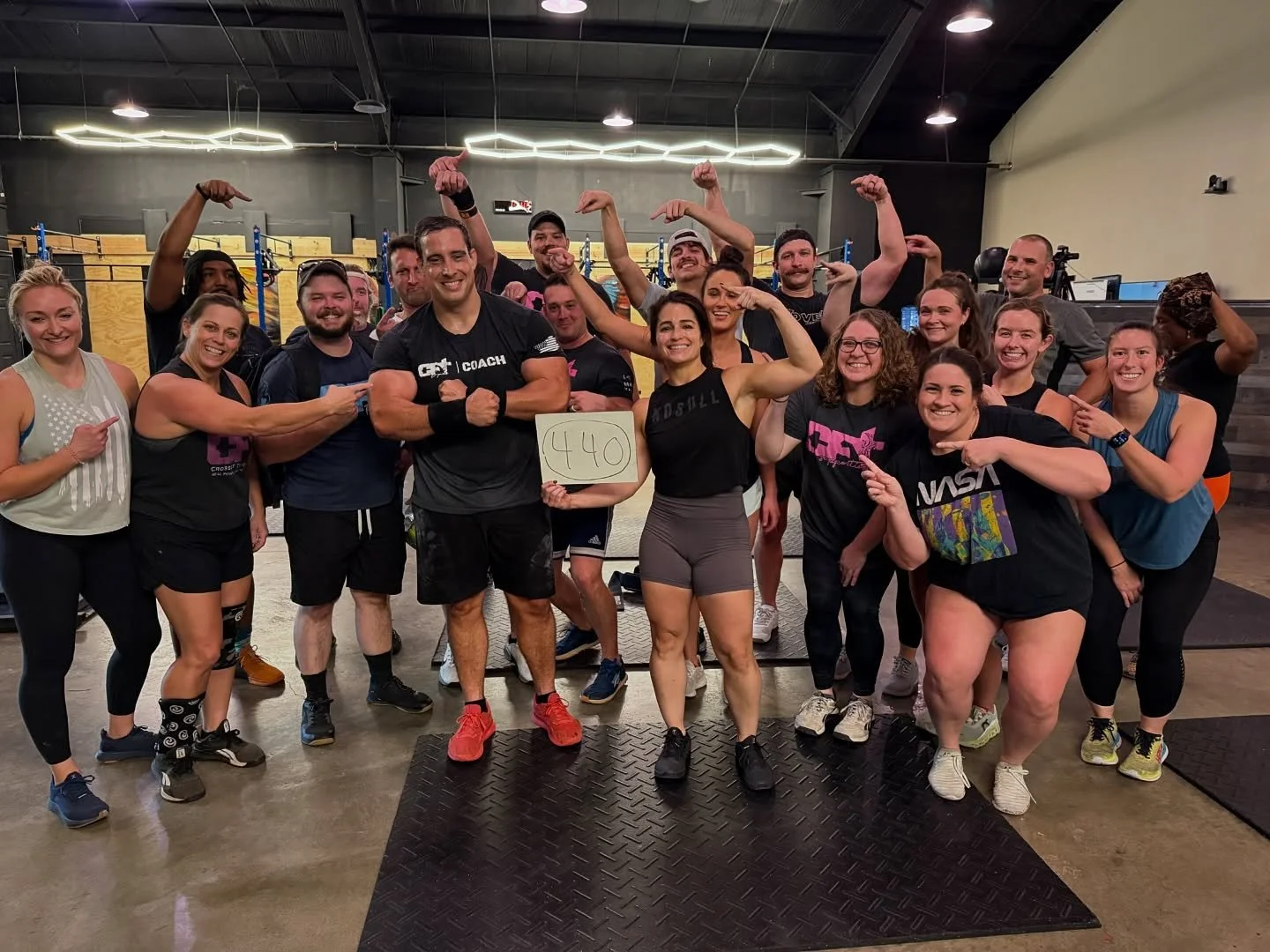 Group of people in gym celebrating, some flexing muscles, holding a sign with '440', smiling, diverse in appearance, gym equipment in background.