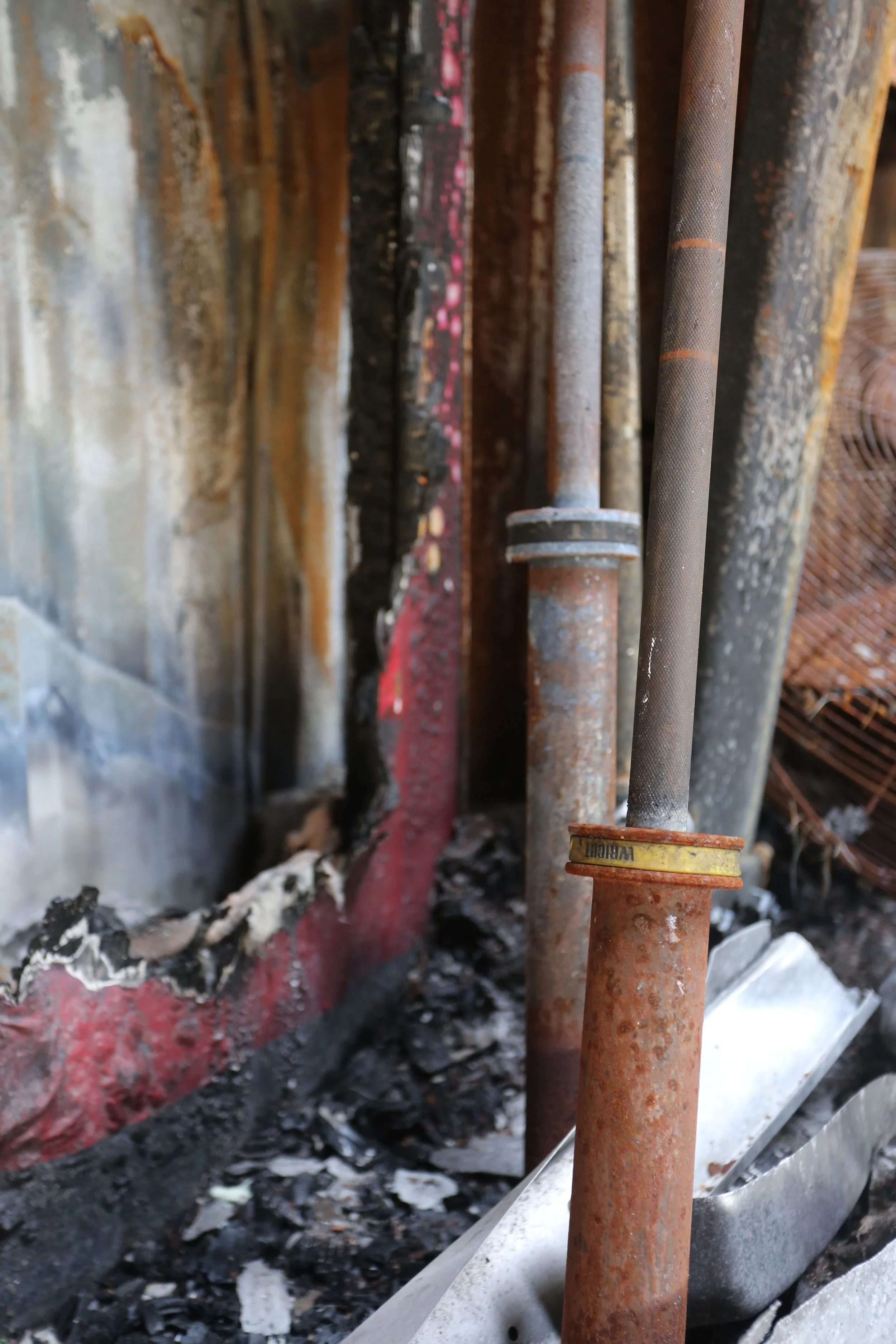 Close-up of rusted pipes and a red-painted metal surface with charred debris and burnt material in the background.