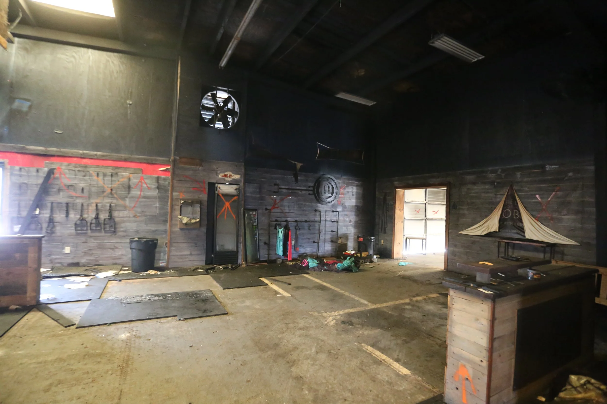Interior of an abandoned or neglected building with blackened walls, a ceiling fan, and spray-painted arrows and markings on the walls. The floor is cluttered with debris, clothing, and trash. There is a small wooden table and some industrial-style s